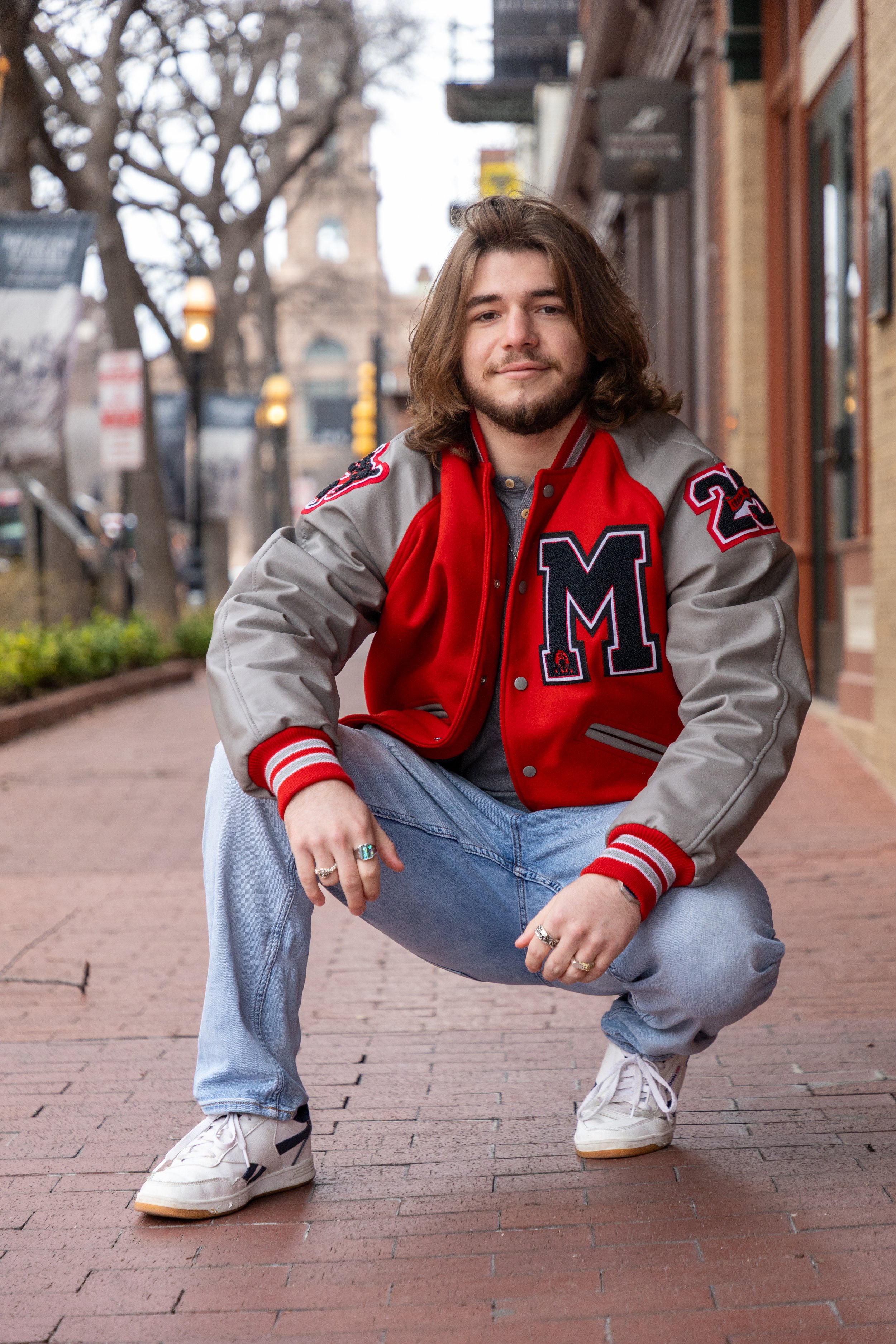 A young man with long hair and a beard is squatting on a brick sidewalk in an urban area. He is wearing a red and gray varsity jacket with a large letter M on the front, light blue jeans, and white sneakers. The background features trees, streetlight