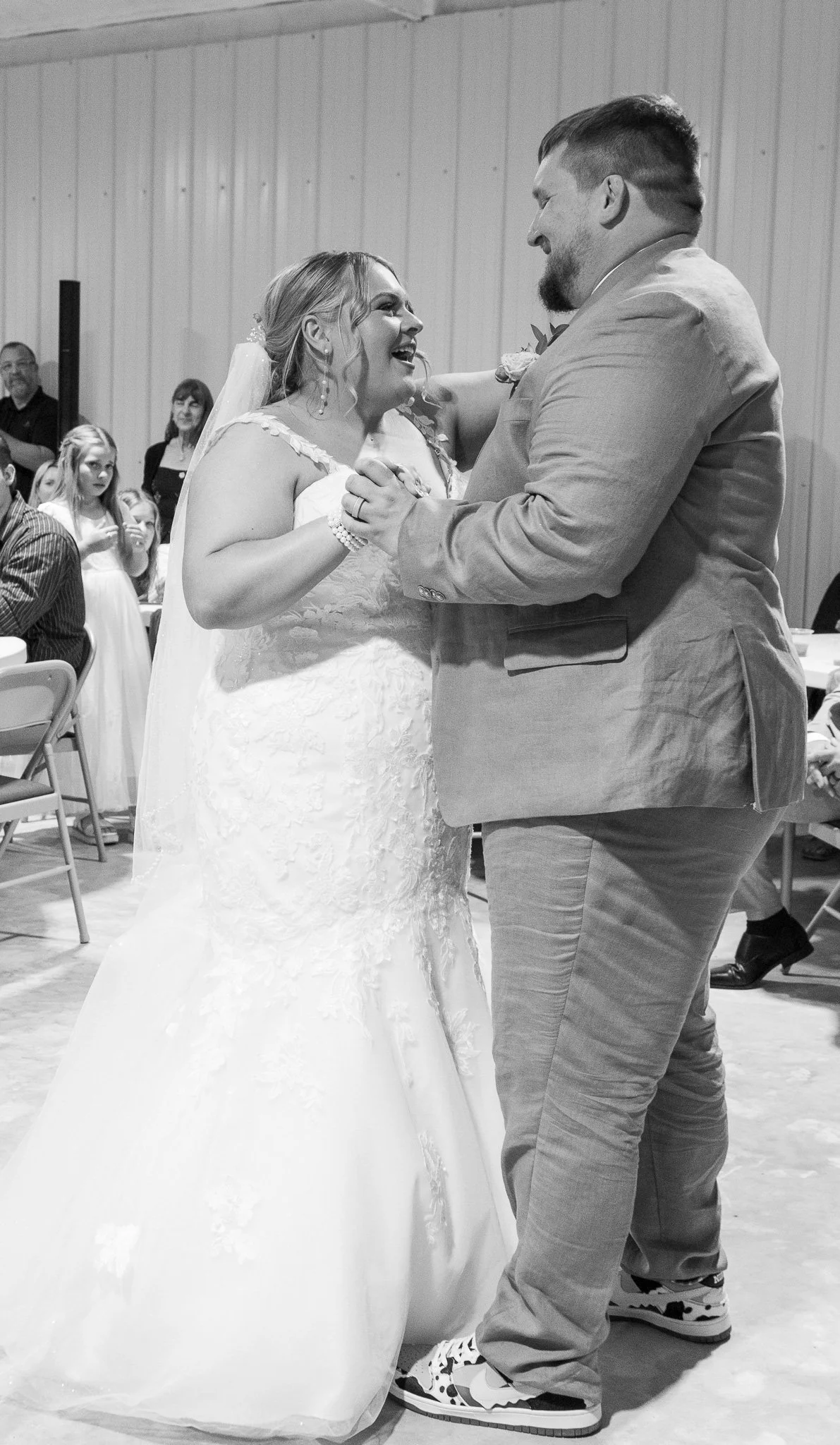 Black and white photo of a bride and groom sharing their first dance at a wedding reception, with guests watching in the background.