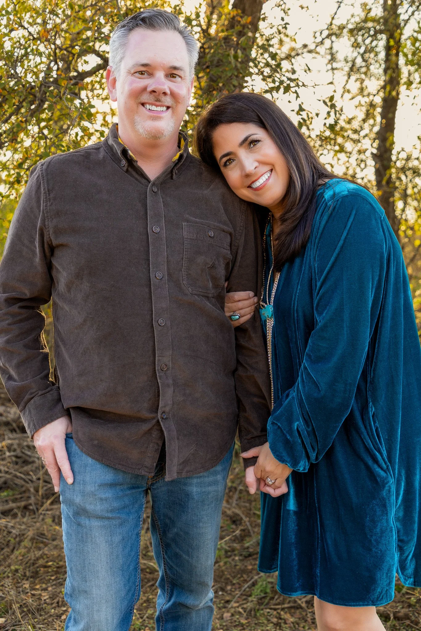 A smiling man and woman holding hands outdoors in a wooded area during golden hour.