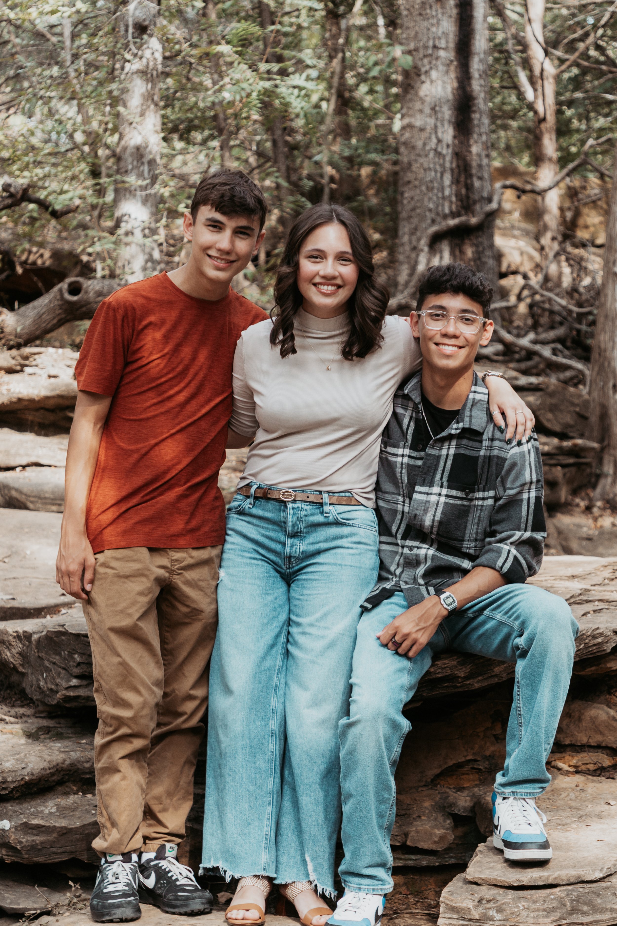 Three young friends smiling, standing outdoors on rocky terrain surrounded by trees.