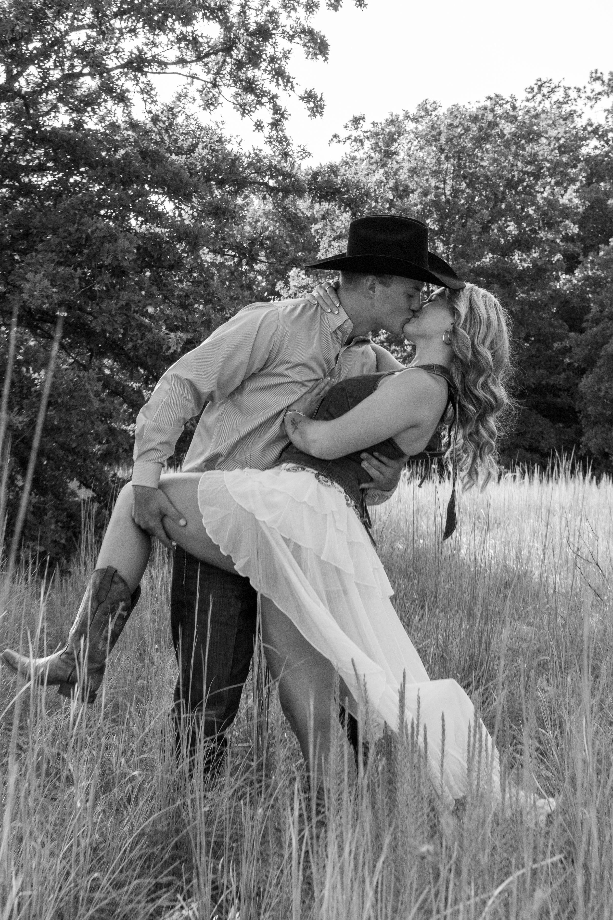 A black-and-white photo of a couple kissing outdoors in a grassy field, with trees in the background. The man is wearing a cowboy hat and the woman has long, curled hair.