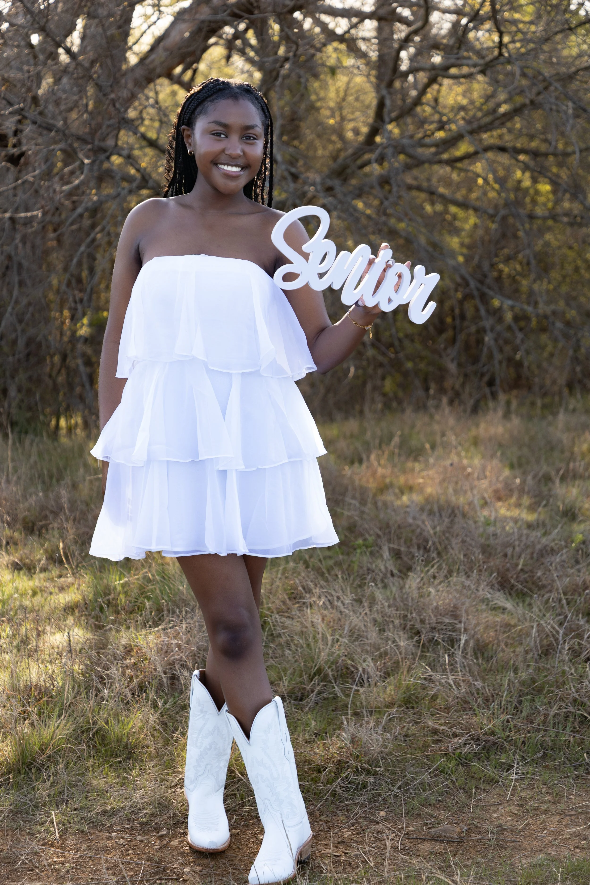 A young woman in a white ruffled dress and white cowboy boots standing outdoors, smiling, holding a sign that says "Senior" amid a background of trees and grass during the day.
