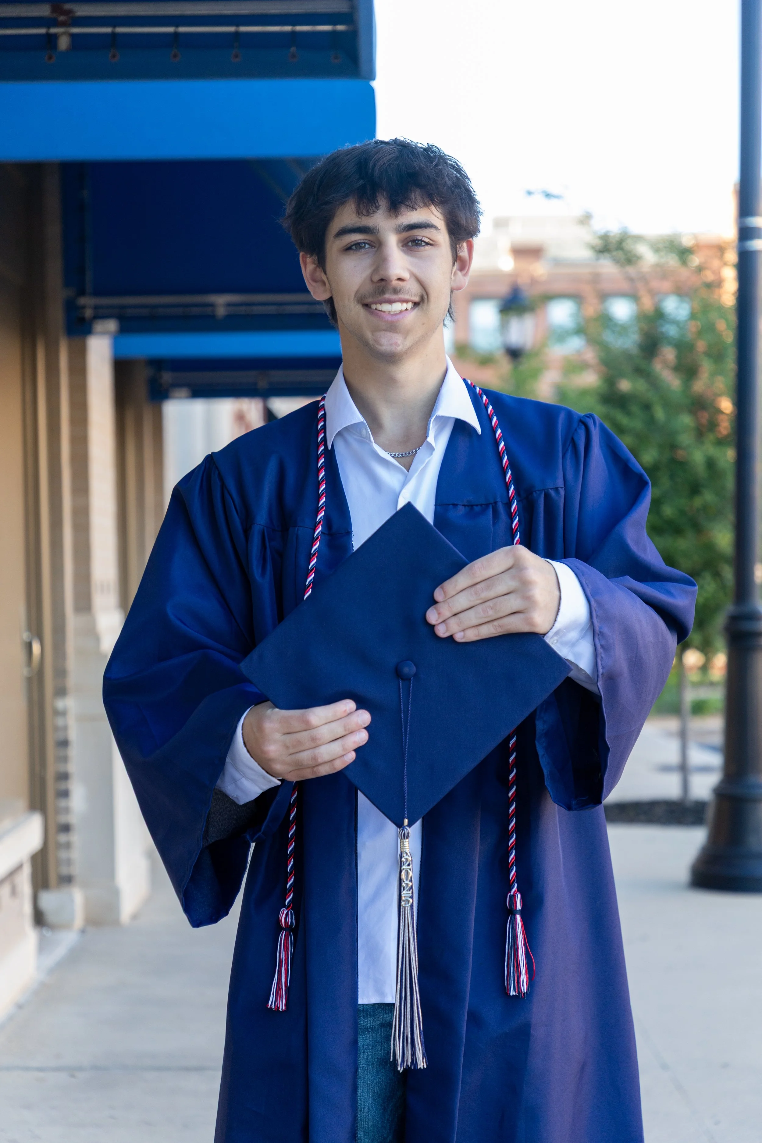 A young man wearing a graduation gown and cap, holding the cap in front of him, standing outdoors on a sidewalk with buildings and trees in the background.