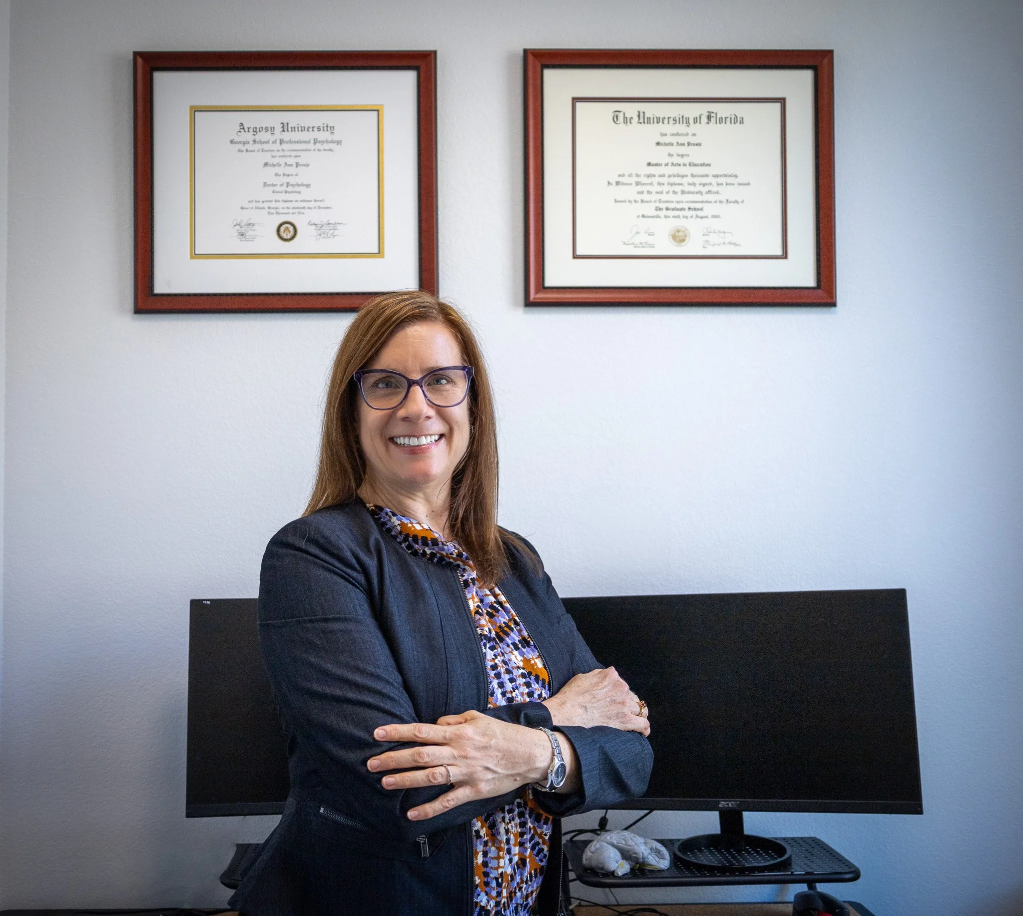 A woman with brown hair, wearing glasses and a dark blazer, standing in an office with two diplomas on the wall behind her. She is smiling with her arms crossed.