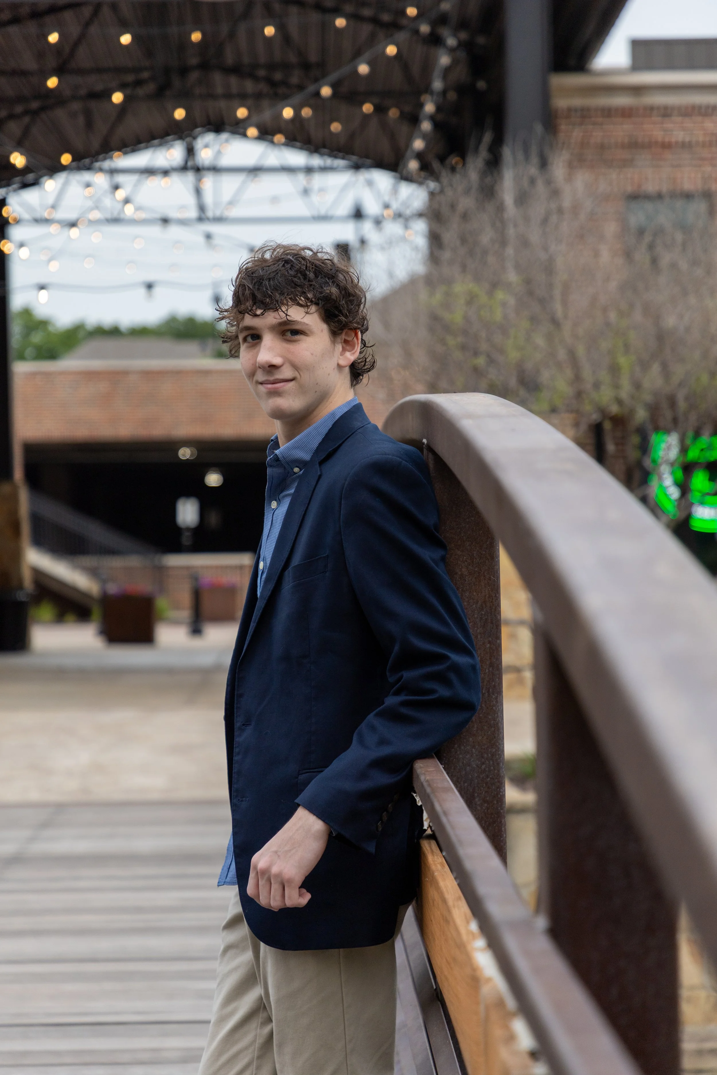 Young man in blue blazer and khaki pants leaning on a wooden and metal railing on a bridge outdoors, with string lights overhead and a brick building in the background.