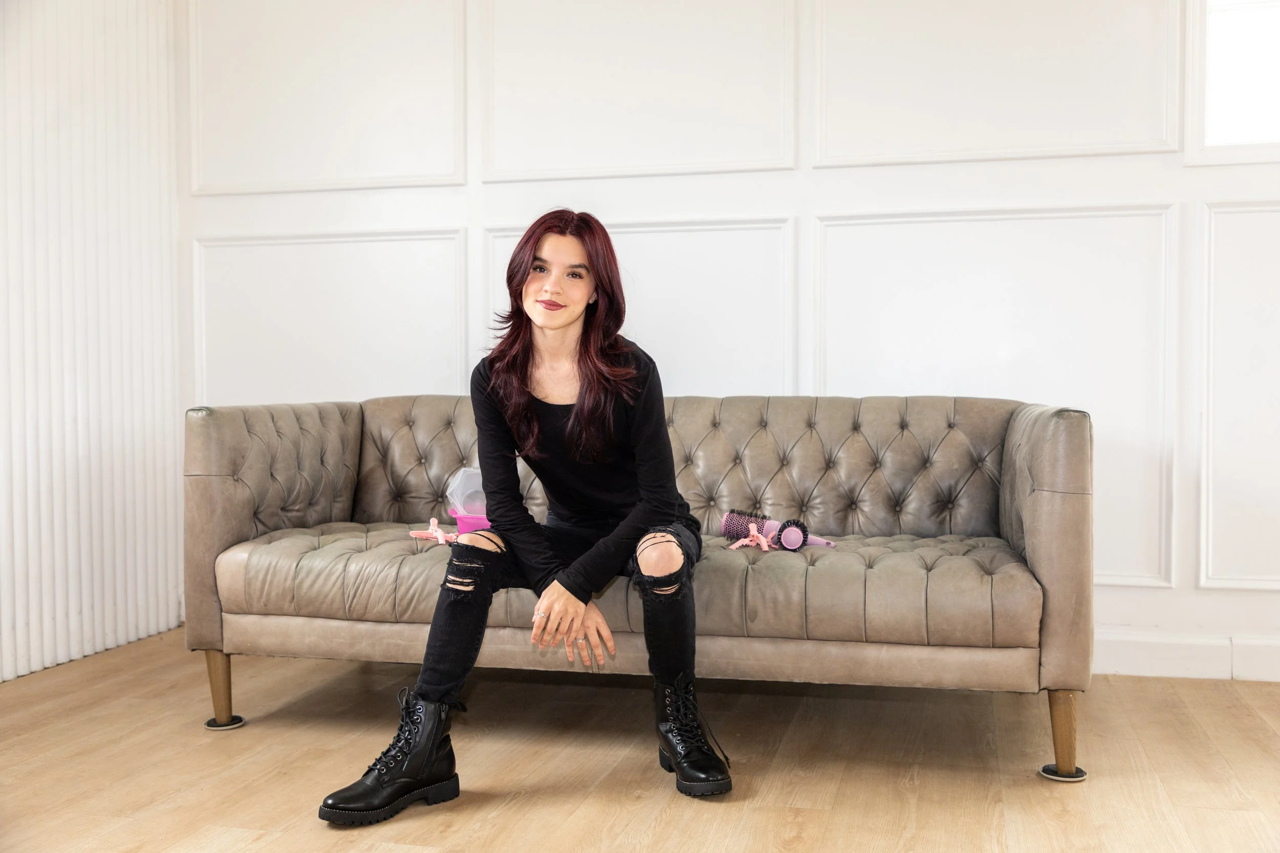 A young woman with long maroon hair, wearing a black long-sleeve shirt, ripped black jeans, and black combat boots, sitting on a beige tufted sofa in a white-paneled room, smiling at the camera with pink hairbrushes and toy accessories on the sofa be