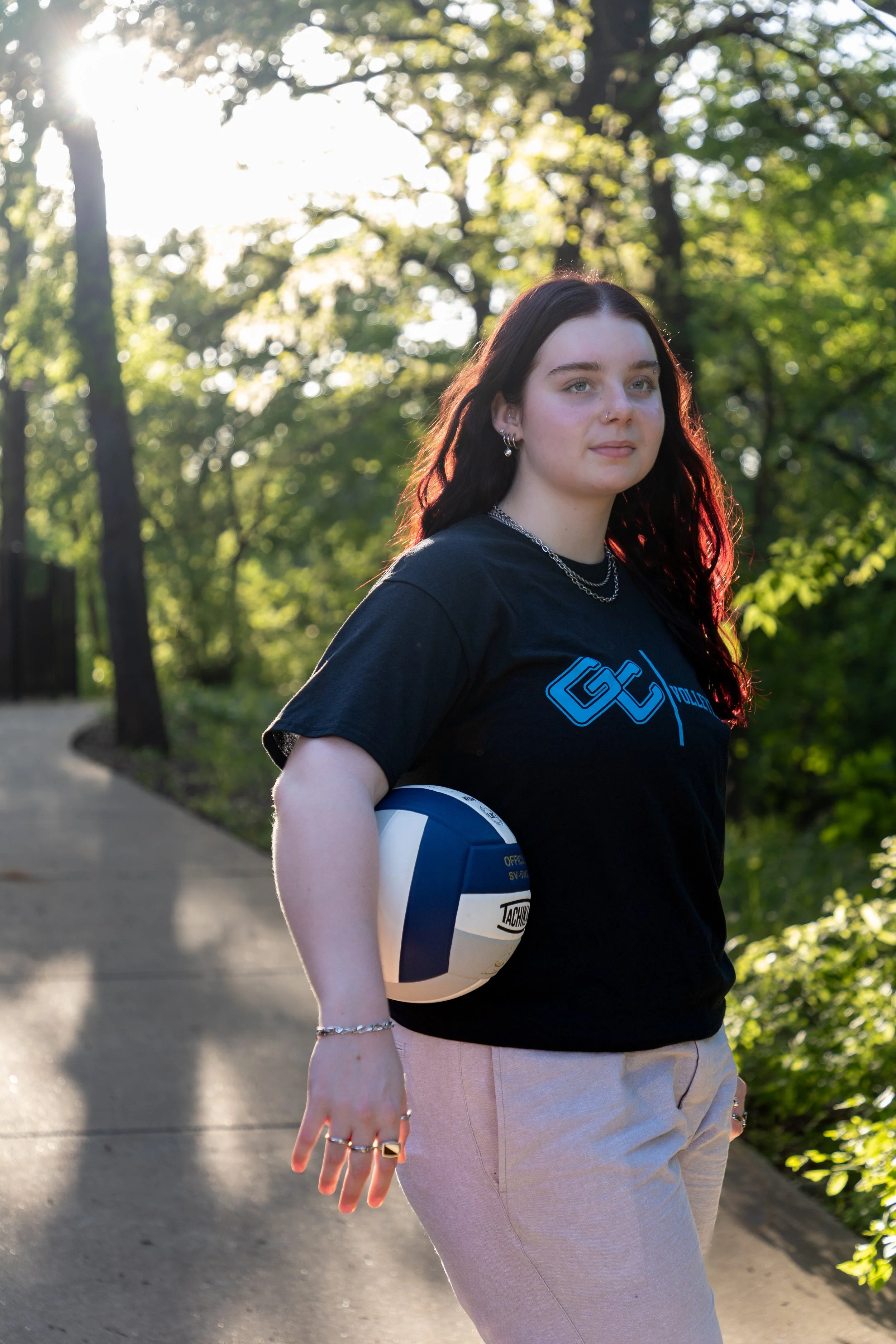 A young woman with dark red hair holding a volleyball in her right arm stands on a shaded park pathway. She wears rings, a bracelet, earrings, and a black t-shirt with blue graphics. The background features green trees and sunlight.