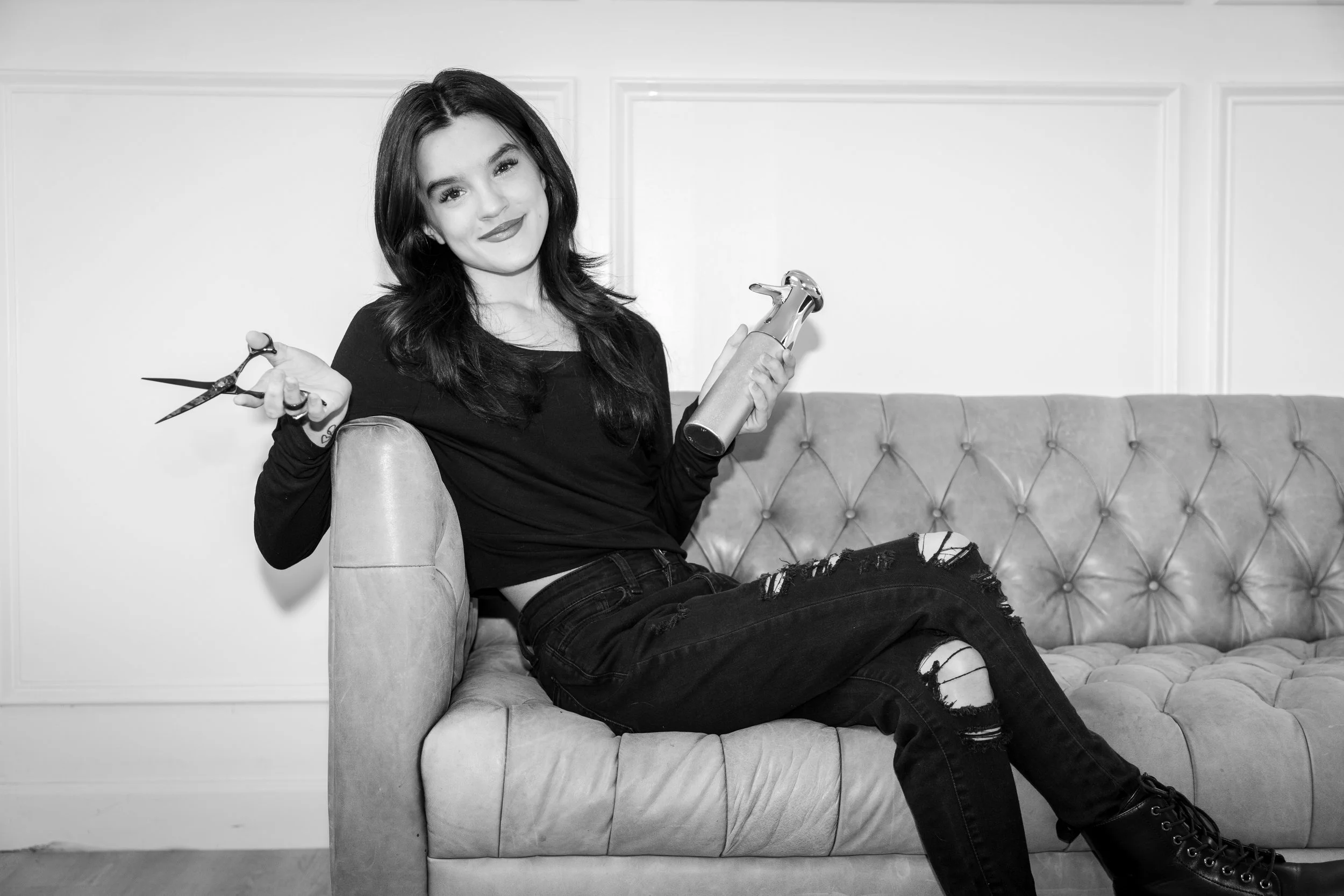 A young woman with dark hair sitting on a vintage sofa, holding hair cutting scissors in one hand and a hairdryer in the other, in a room with paneled walls.