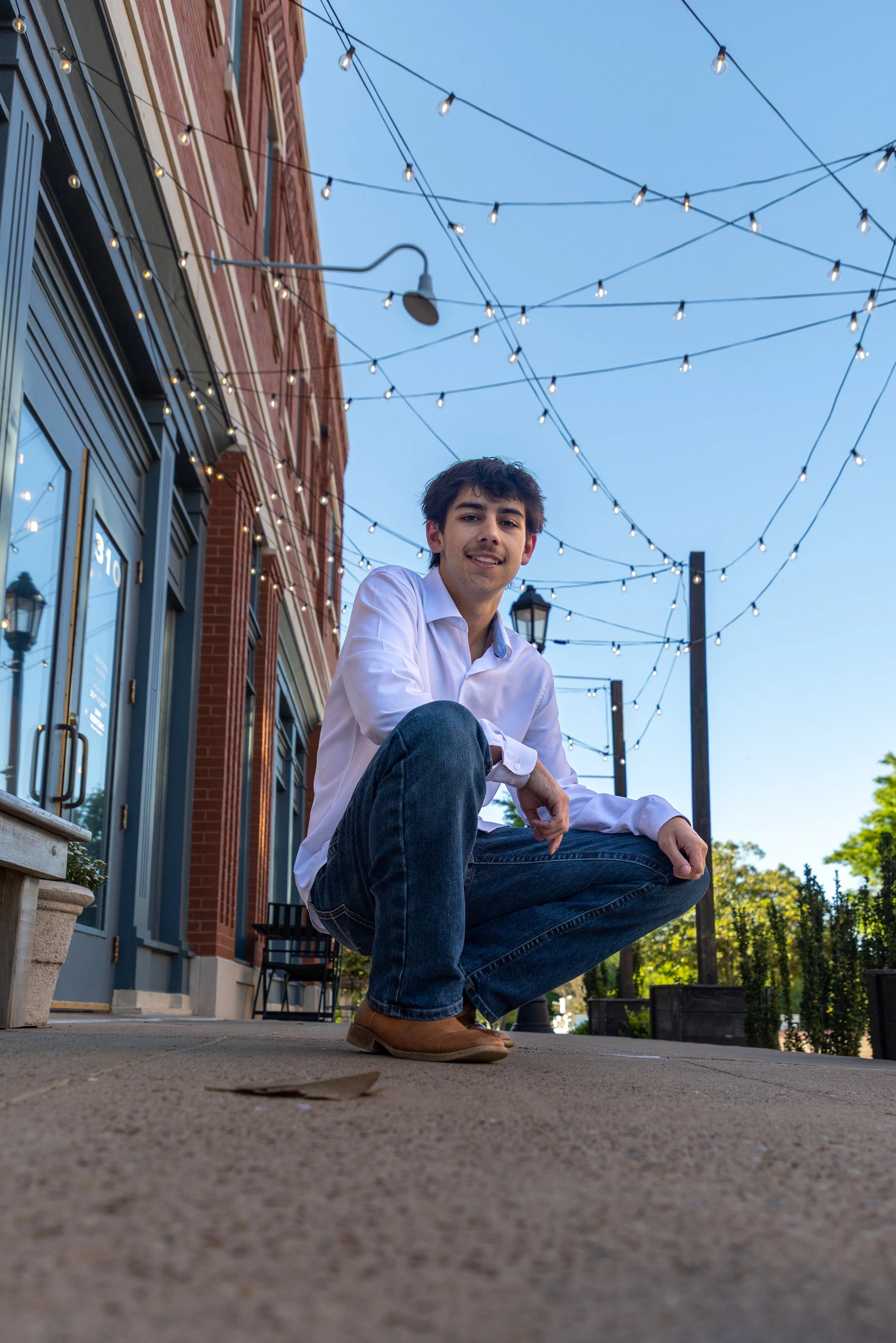 Young man squatting on sidewalk in an outdoor urban setting with string lights, brick building, and trees in the background.