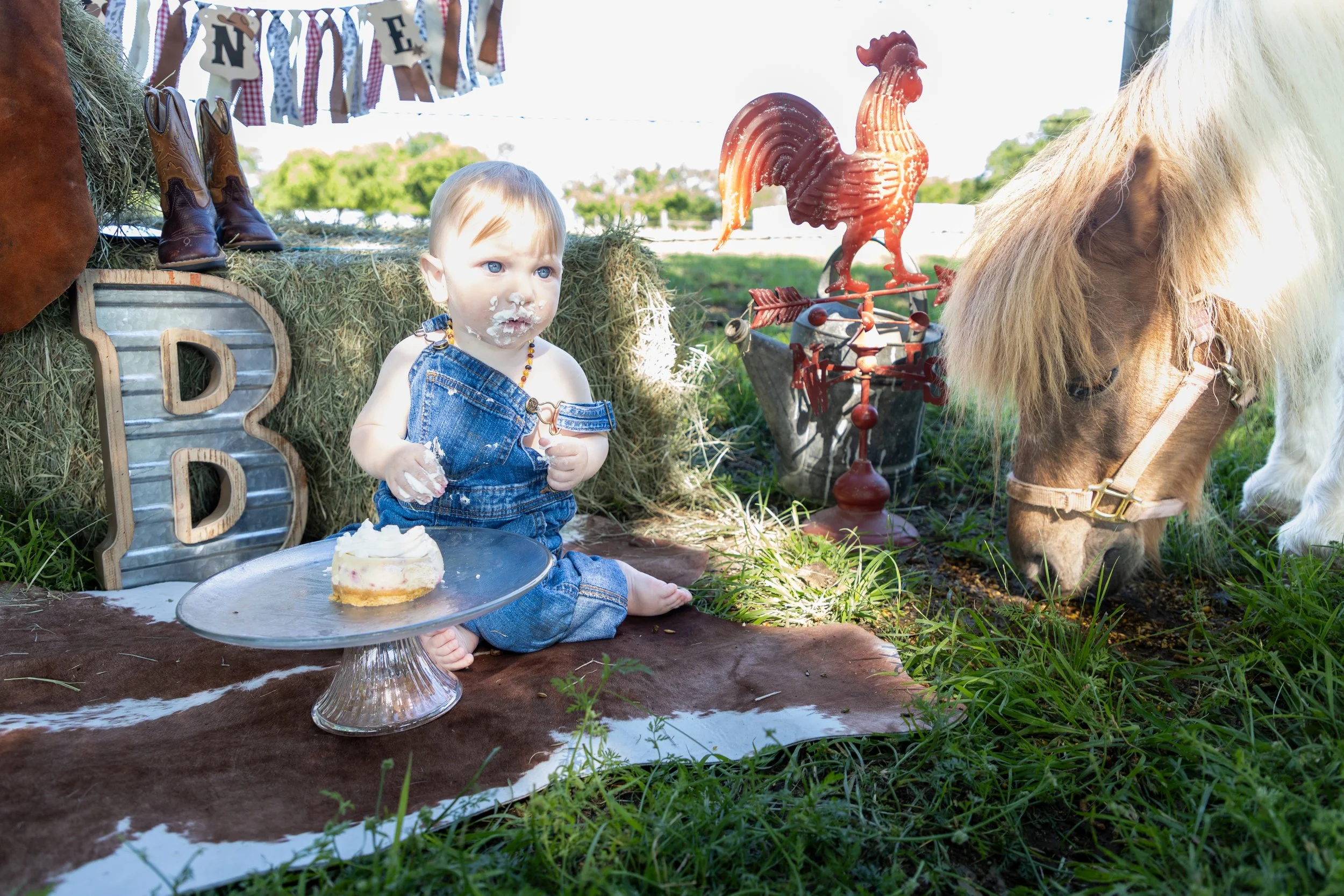 Young child with blue eyes and messy face sitting on a brown animal hide, with a small cake on a silver tray, in a rural outdoor setting. The child is wearing denim overalls and a beaded necklace. There is a large hay bale behind the child, decorated