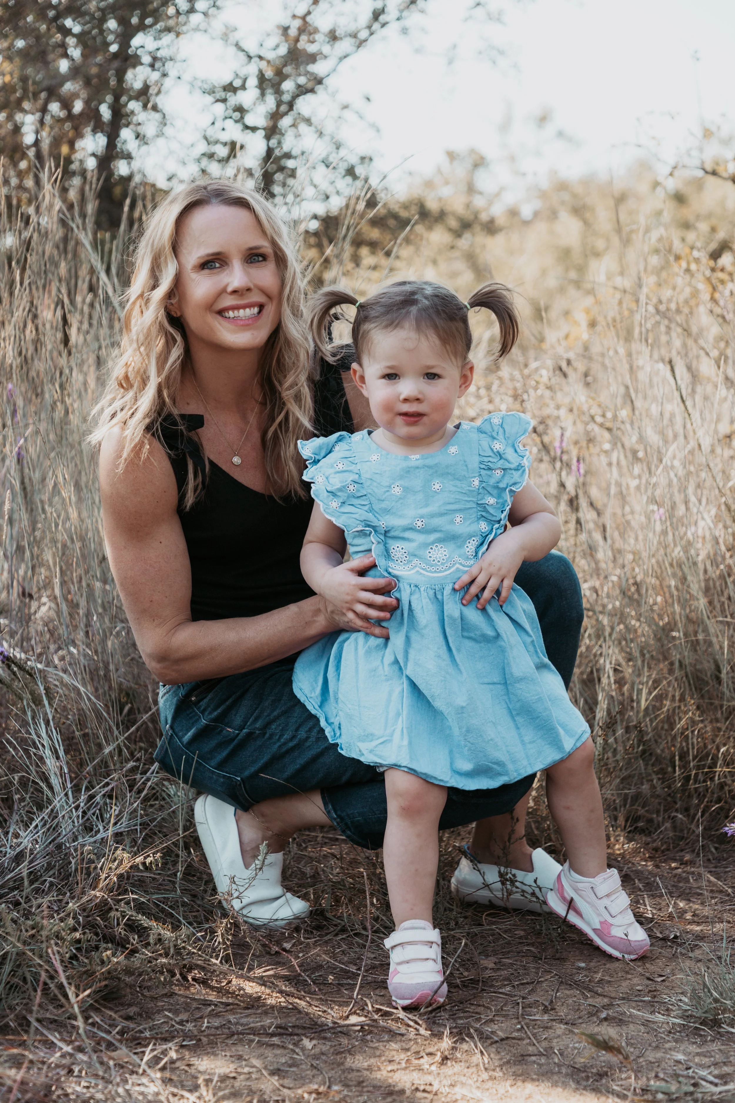 A woman with blonde, wavy hair and a young girl with pigtails and a blue dress sit outdoors in a field of tallDry grass with trees in the background. The woman is smiling at the camera while holding the girl, who is looking slightly to the side with 