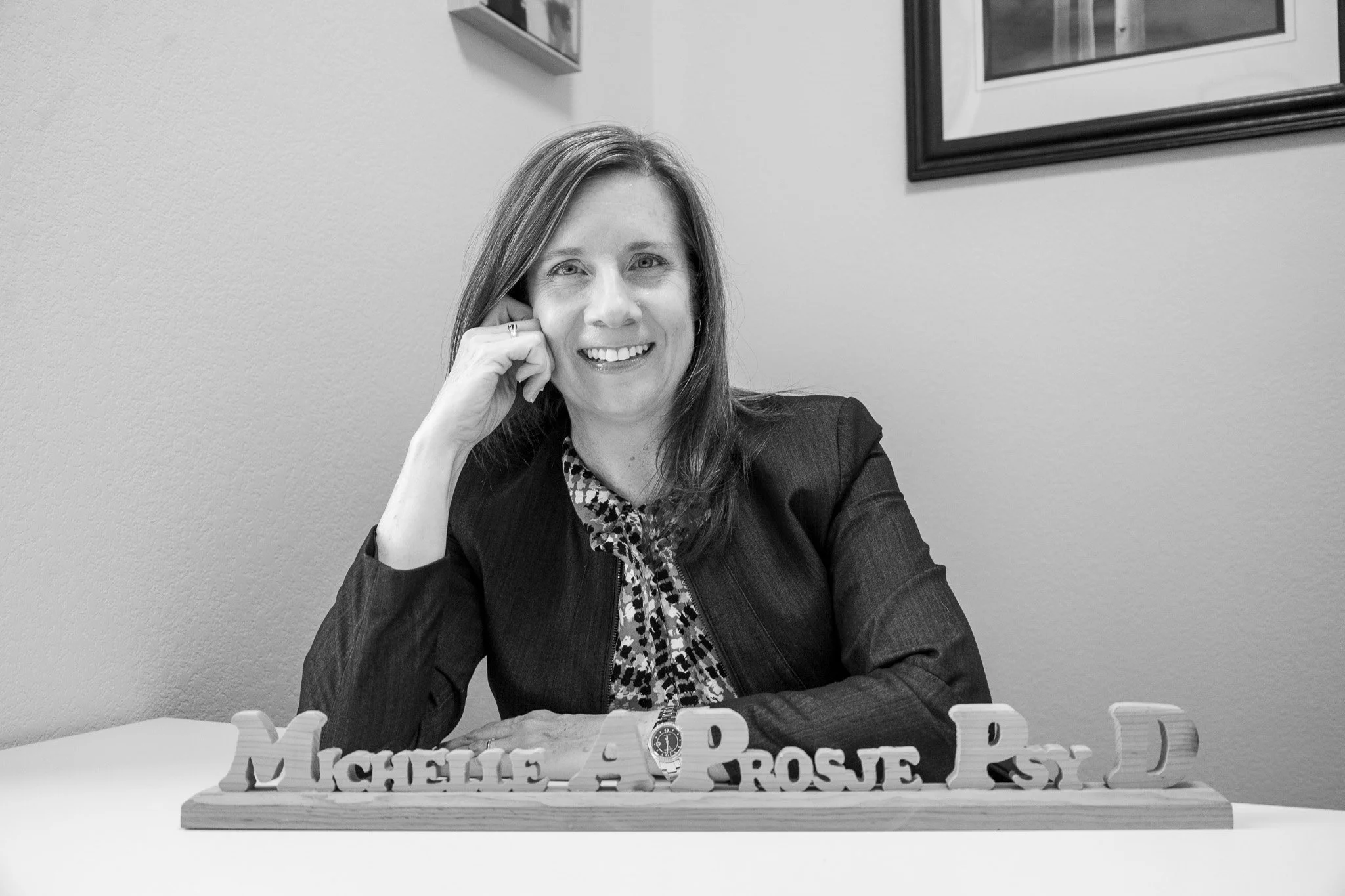 A woman with shoulder-length hair, wearing a dark blazer and patterned blouse, sitting at a table, smiling, with her left hand resting on her face, and a wooden nameplate in front of her that reads "Michelle A. Rose P. D."