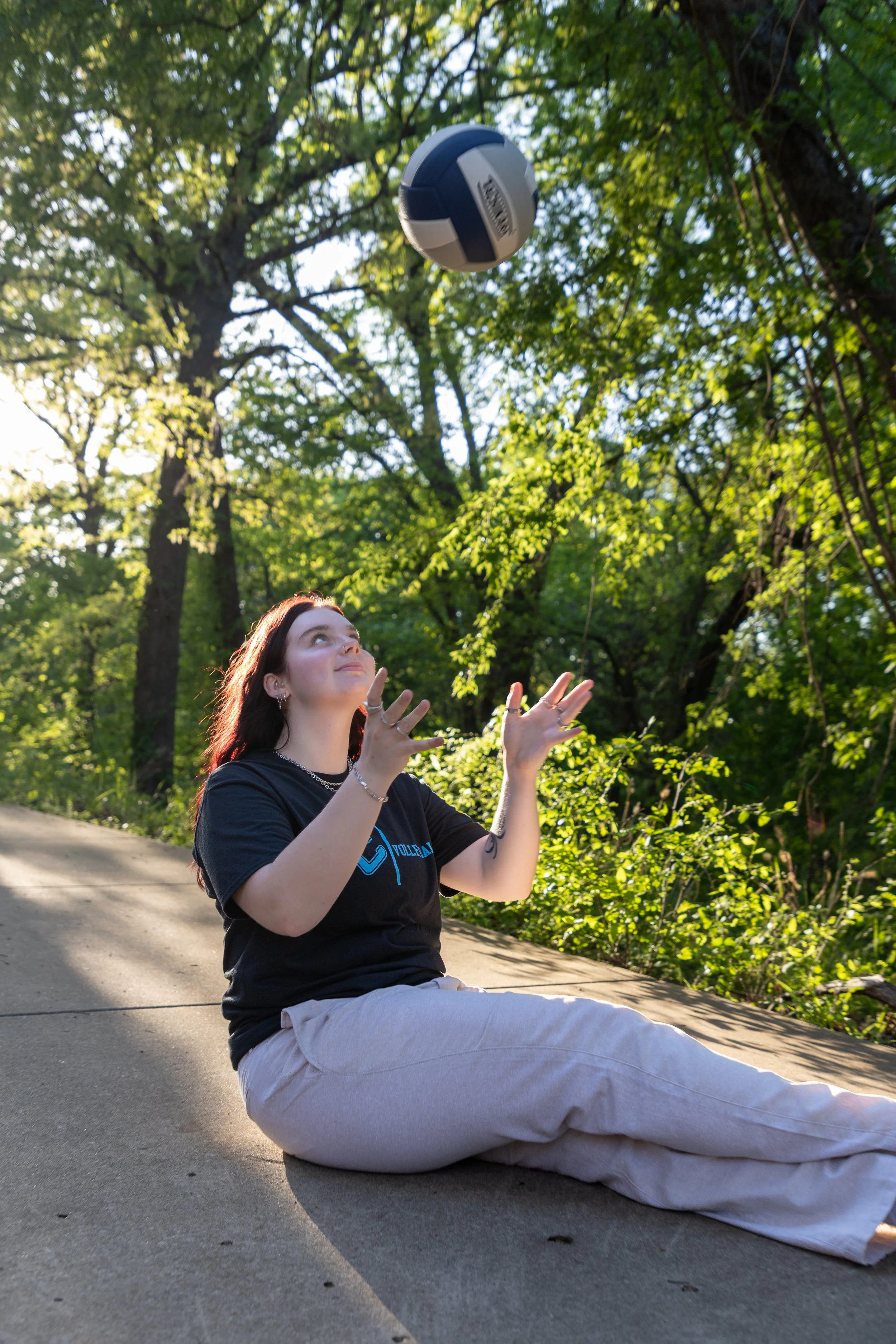 A young woman with red hair sitting on a concrete sidewalk outdoors, playing with a volleyball in the air, surrounded by green trees and sunlight.