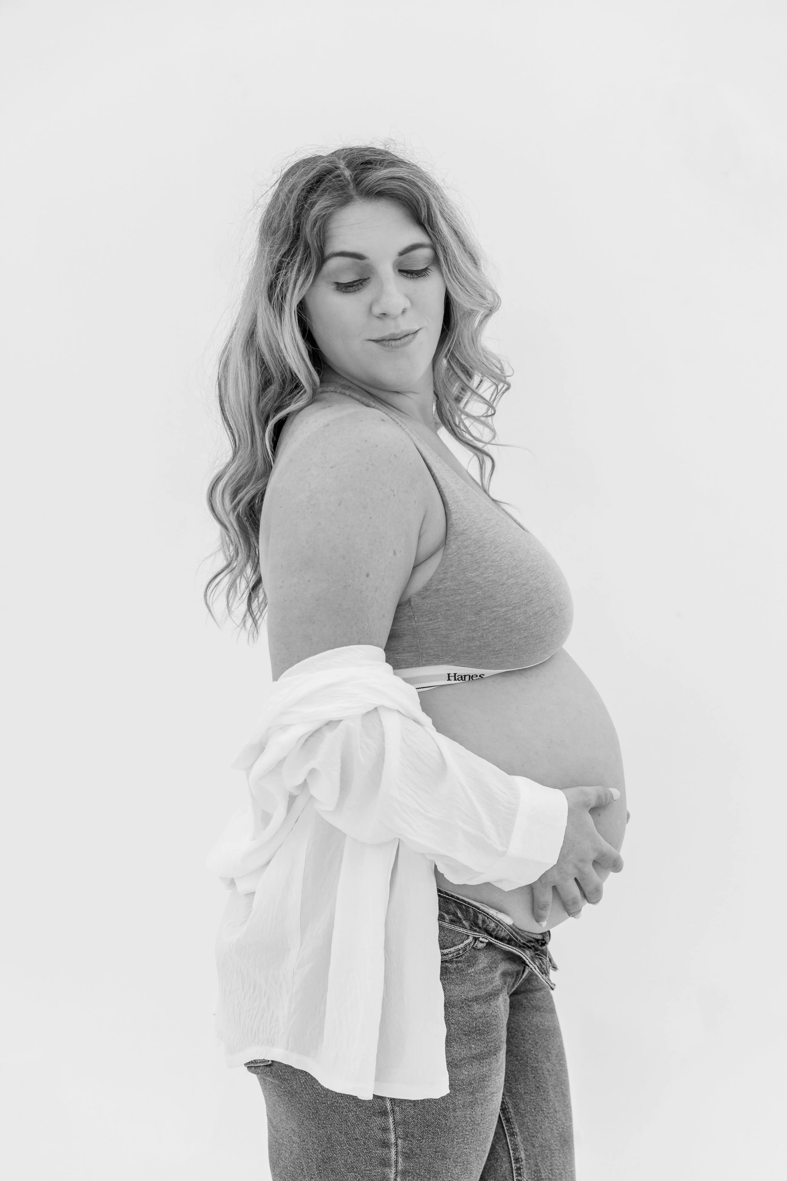 Black and white photo of a pregnant woman with wavy hair wearing a tank top and denim jeans, a white shirt draped over her arm, and another person partly visible behind her.