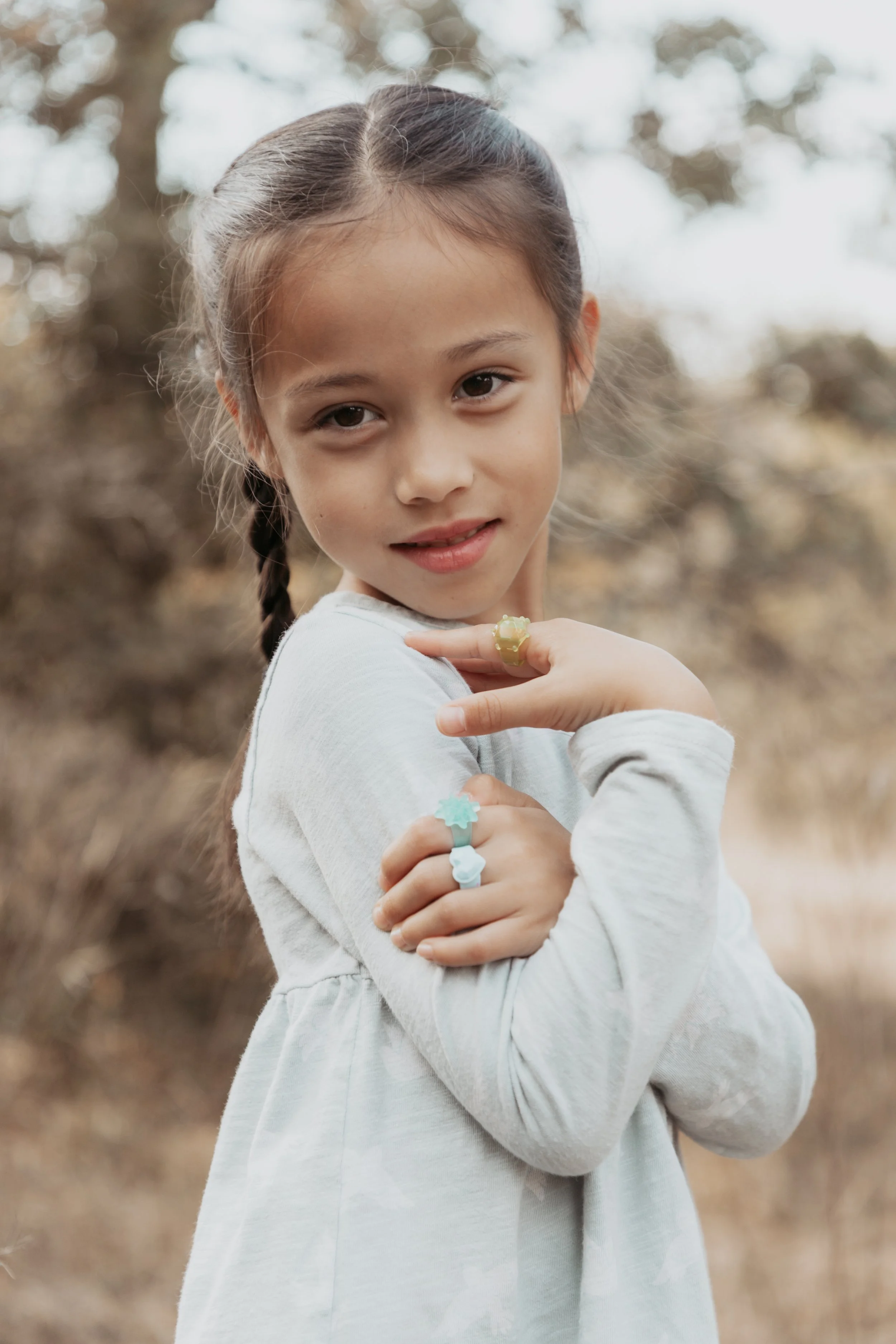 A young girl with braided hair and light skin, smiling outdoors, wearing a white long-sleeve dress and colorful rings, with a blurred natural background.