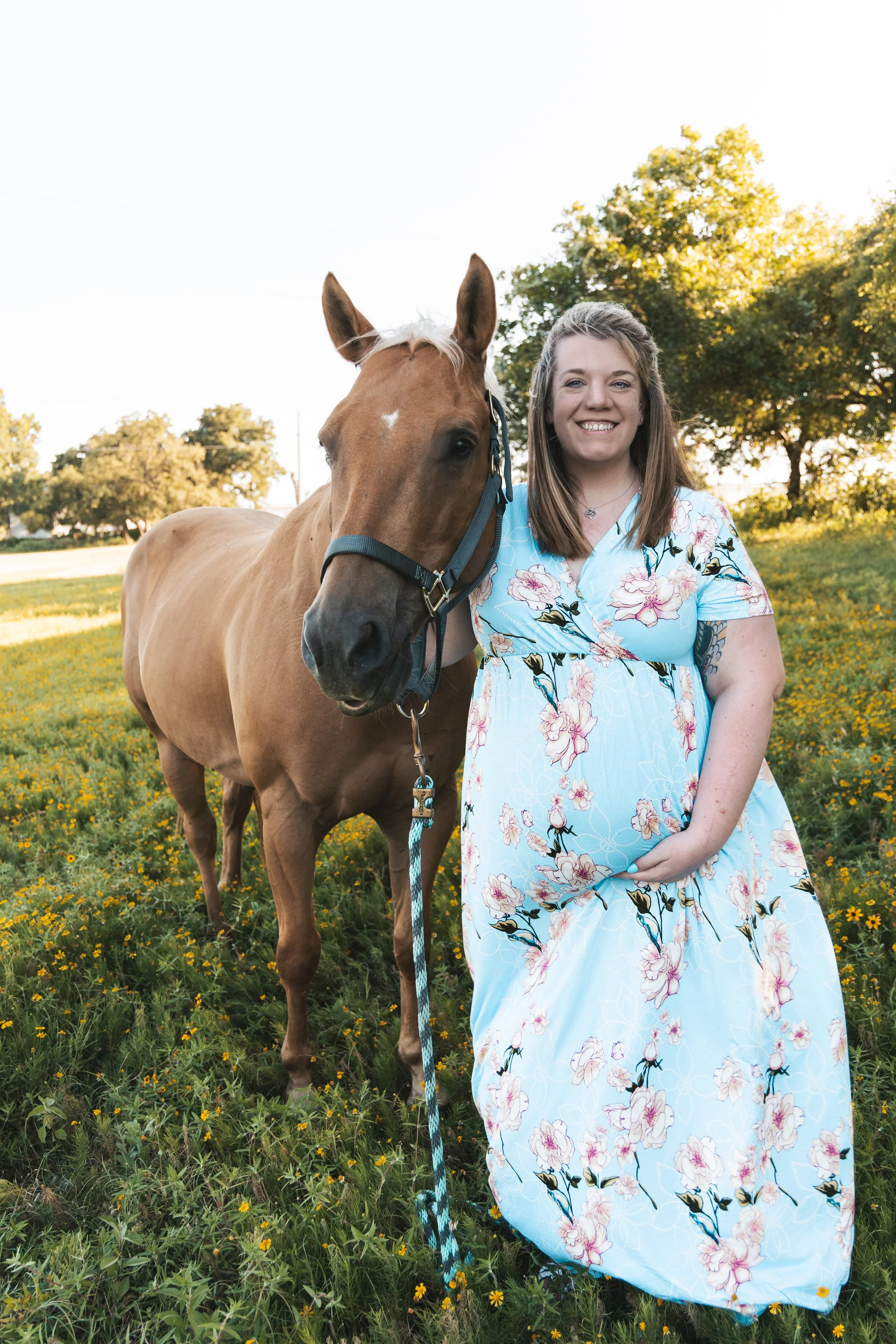A young woman in a floral dress smiling standing next to a chestnut horse with a white blaze in a field of yellow flowers and trees during sunset.