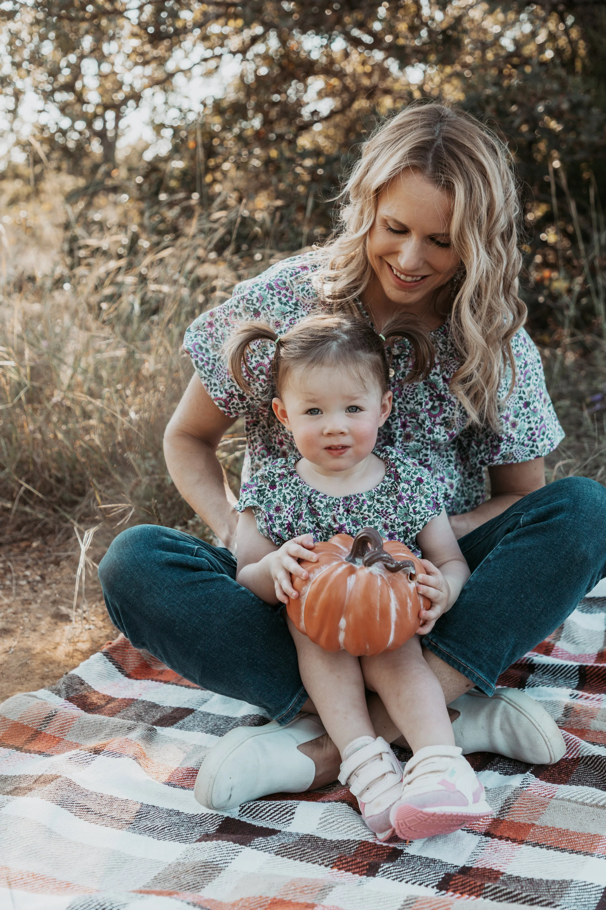 A woman with curly blonde hair and a young girl with pigtails sitting on a plaid blanket outdoors, holding a small pumpkin. The woman is smiling and looking down at the girl, who is holding the pumpkin, with a background of trees and autumn foliage.