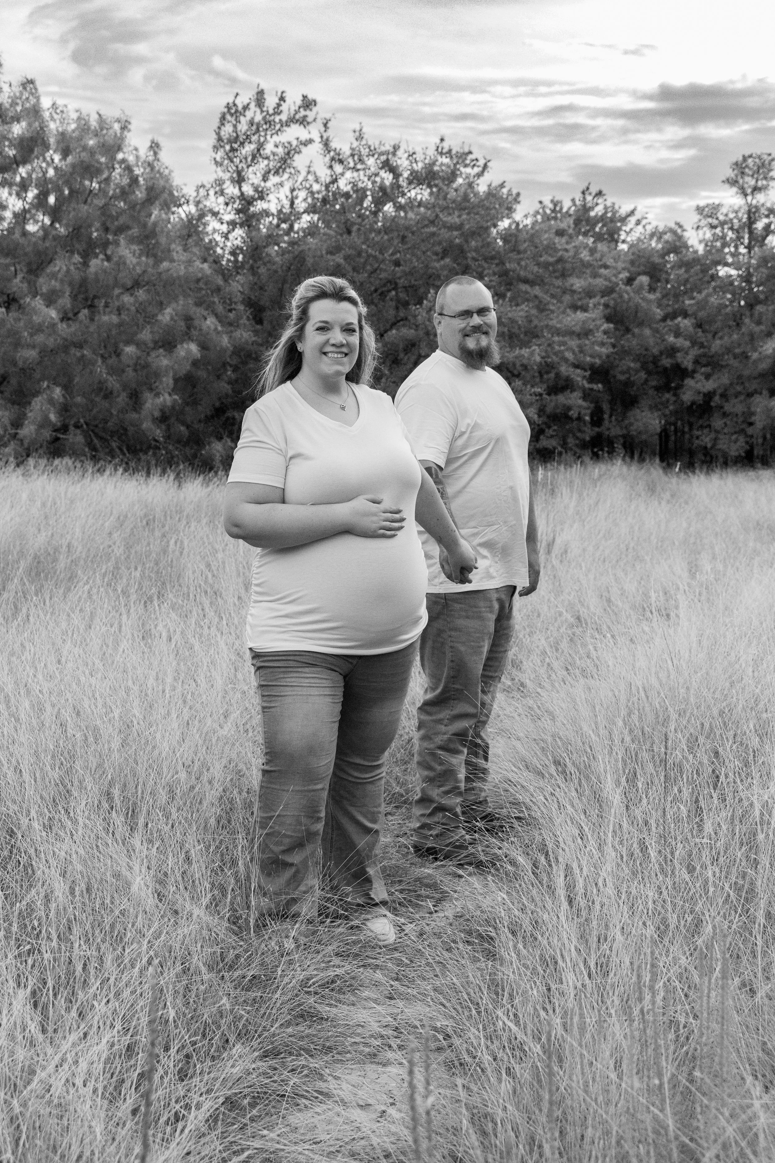 A pregnant woman and a man holding hands in a grassy field with trees in the background, smiling at the camera, in black and white.