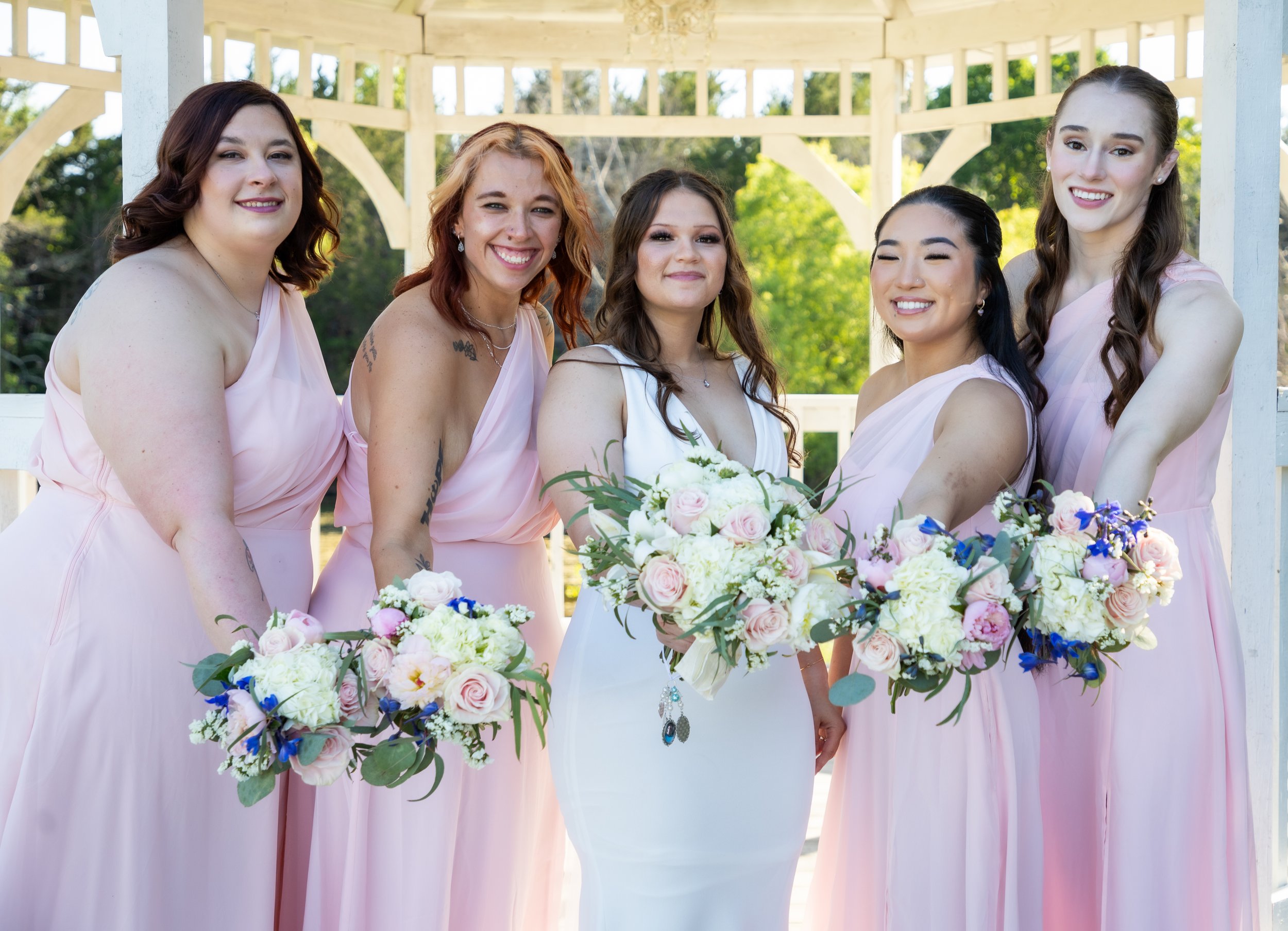 Group of five women in pink dresses, holding bouquets, smiling outdoors during a wedding or celebration.