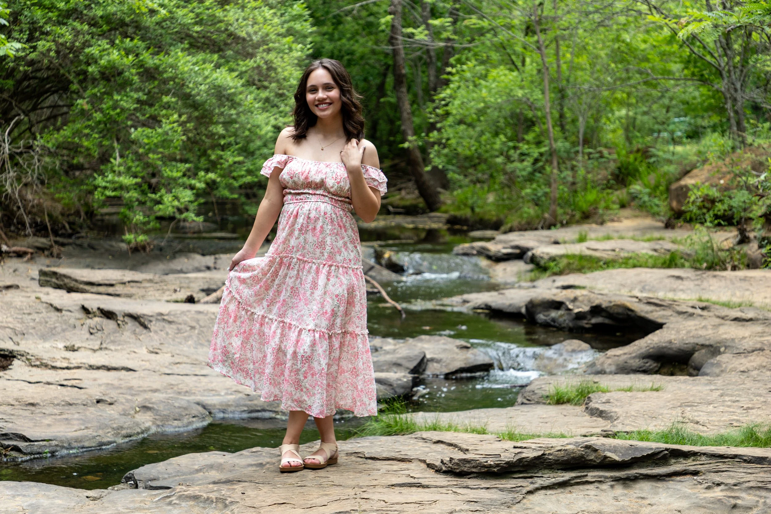 A woman in a pink floral summer dress standing on rocks in a creek surrounded by green trees.