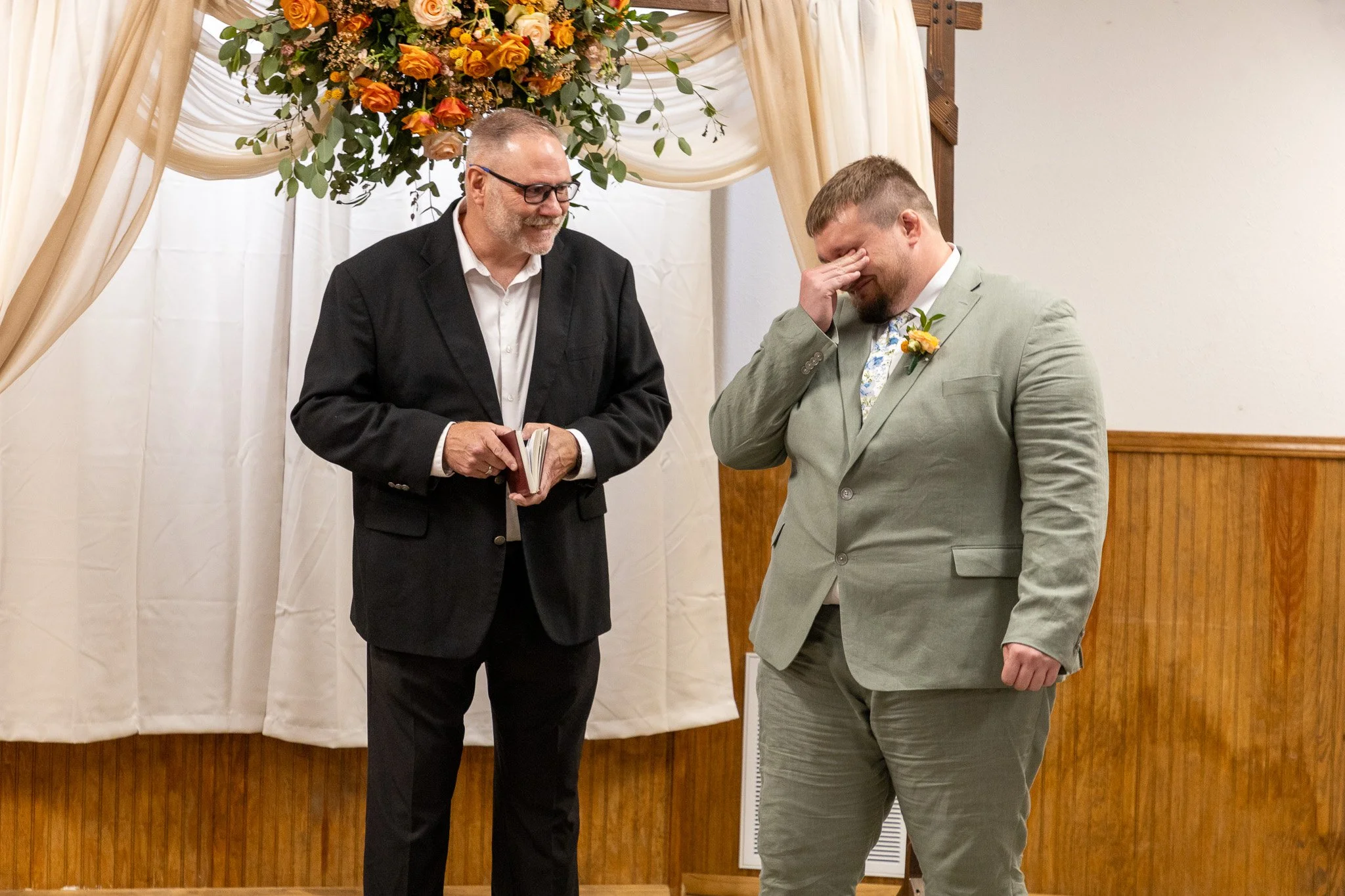 A groom in a light gray suit, with a yellow boutonniere, standing with his hand covering his face, as he looks down, while a man in a black blazer and white shirt, holding a book, looks at him and smiles during a wedding ceremony. There is a floral a