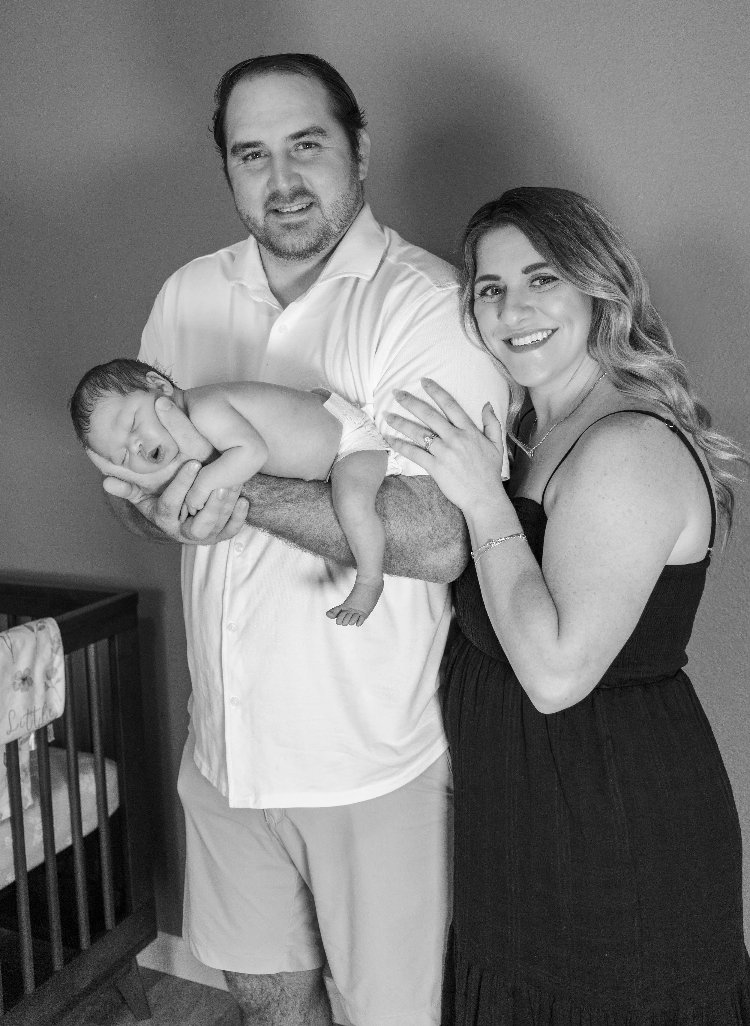 A black and white photo of a couple holding a sleeping newborn baby in a home nursery, with a crib and baby blanket visible in the background.