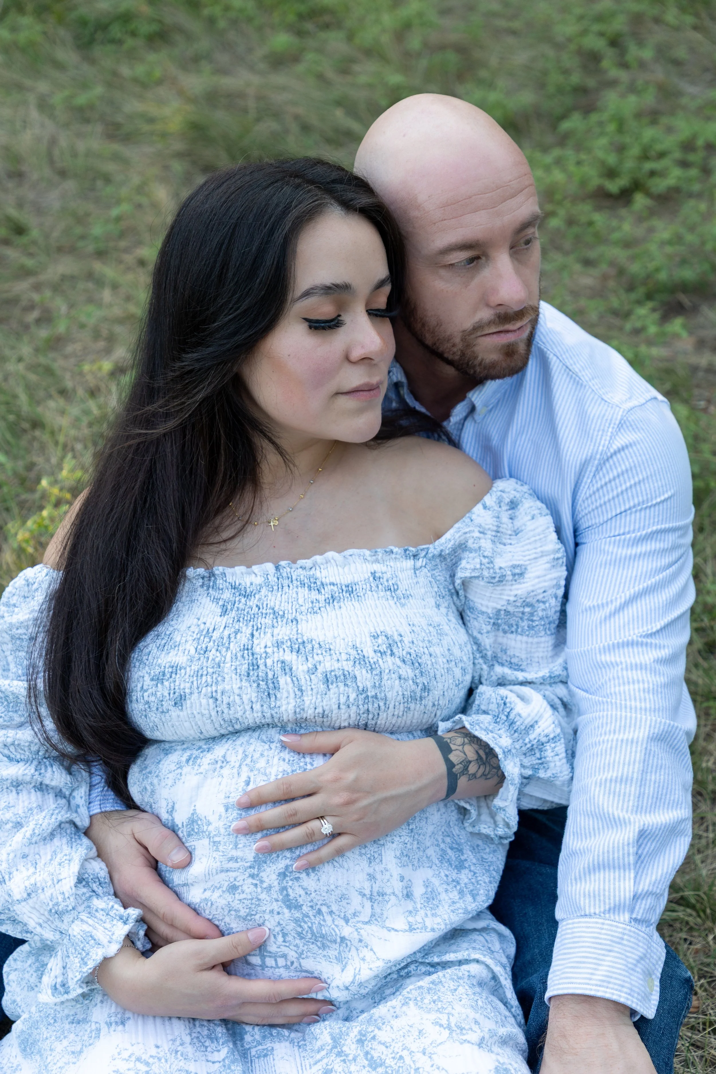 A couple sitting on grass, embracing with their eyes closed. The woman has long dark hair and is wearing a light blue, off-the-shoulder dress. The man is bald with a beard, wearing a light blue striped shirt. The woman is showing her engagement ring.