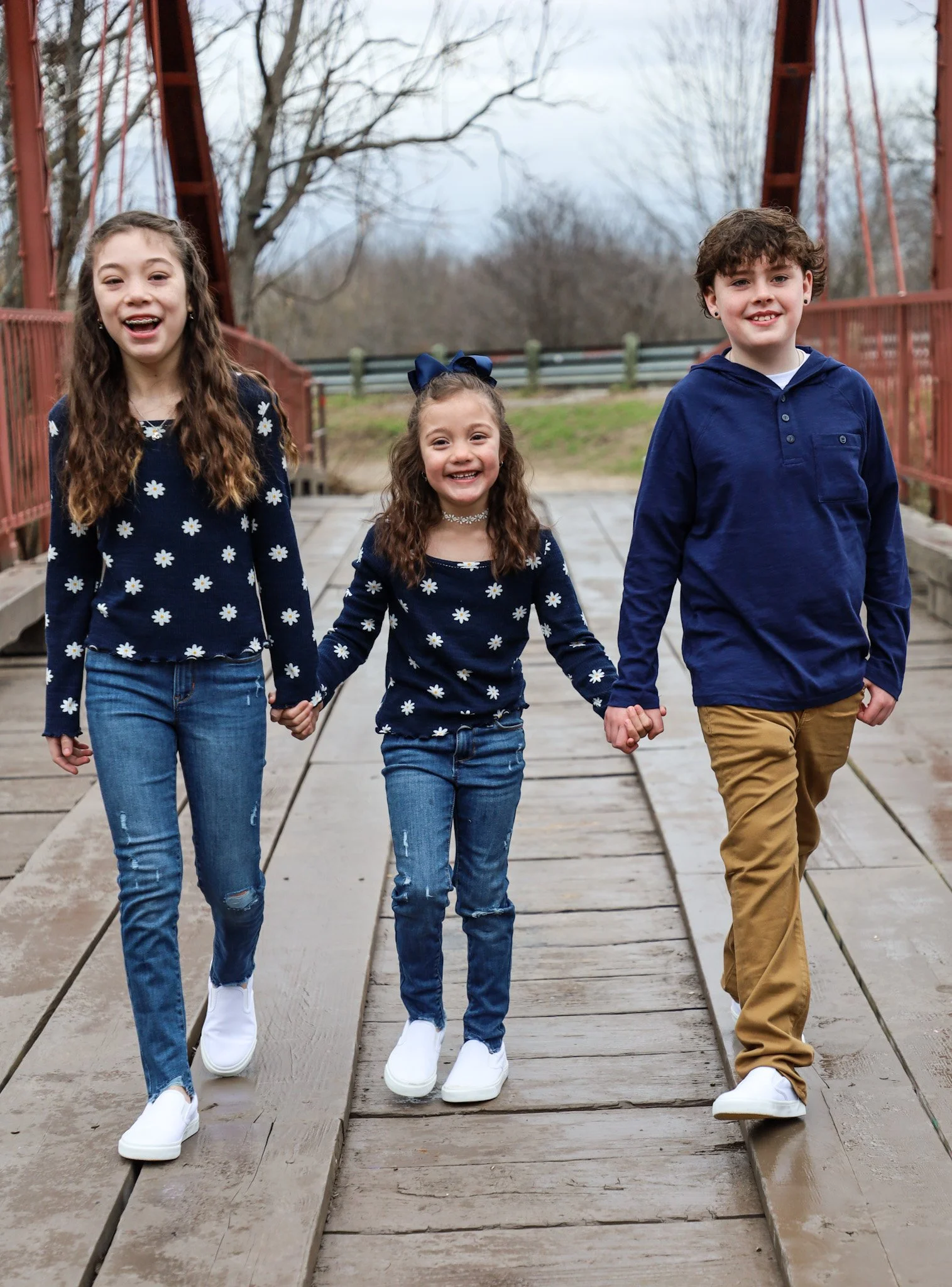 Three children holding hands and walking on a wooden bridge outdoors, wearing matching navy blue sweaters with white daisy patterns, surrounded by trees and a cloudy sky.