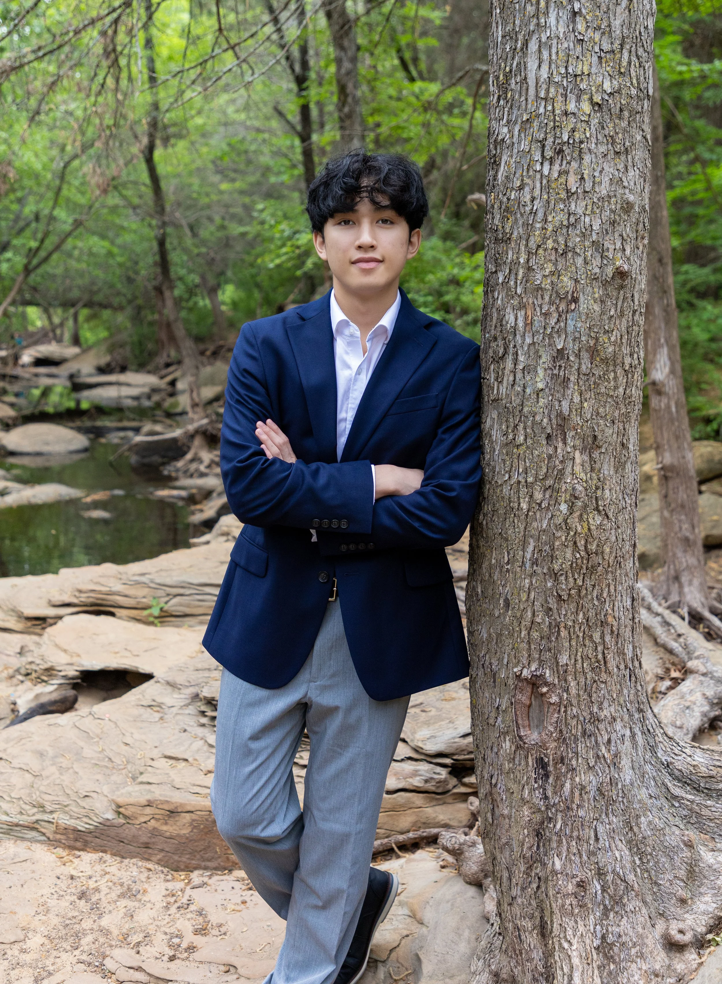 A young man in a navy blazer and light gray trousers standing outdoors near a tree with arms crossed, in a natural setting with rocks, a stream, and green foliage.