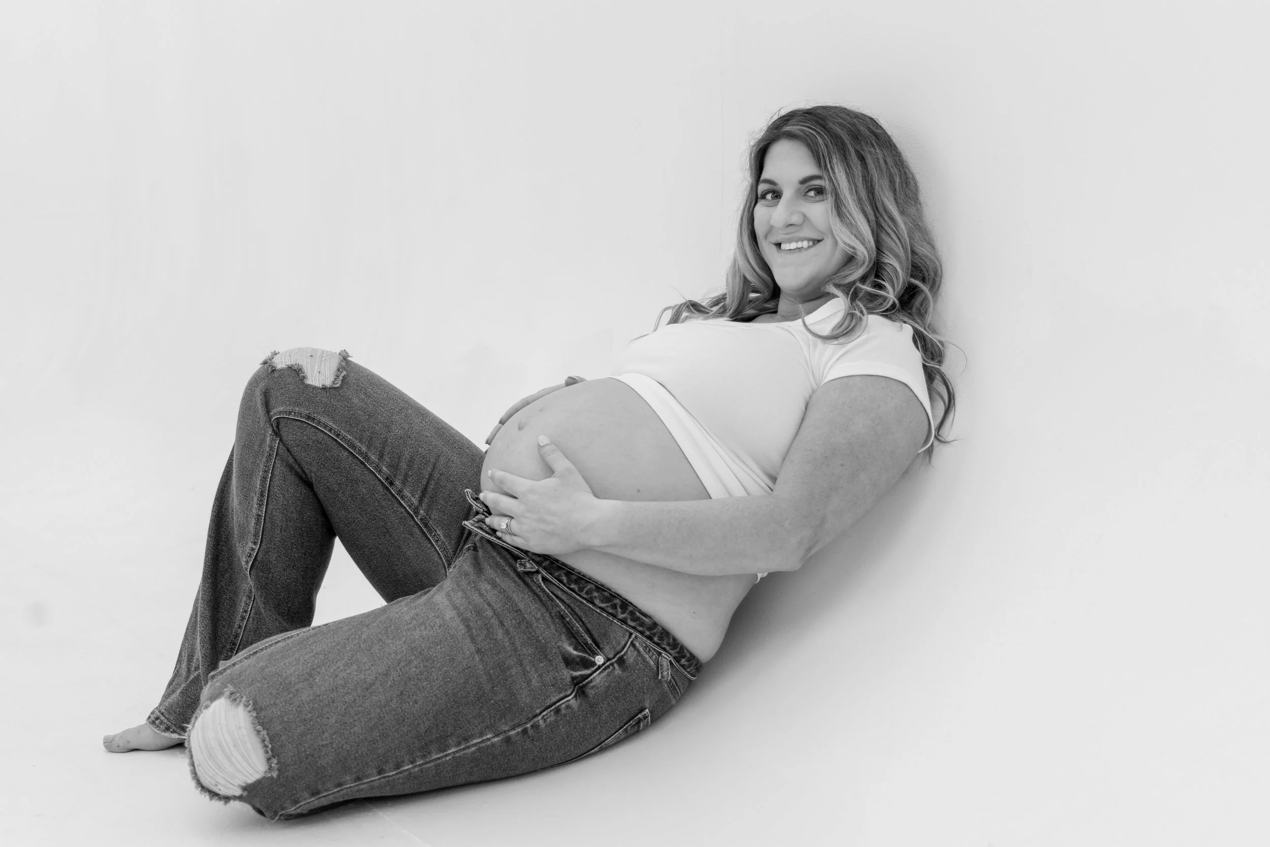 Black and white photograph of a pregnant woman sitting on the floor with her back against a plain wall, smiling and holding her belly.