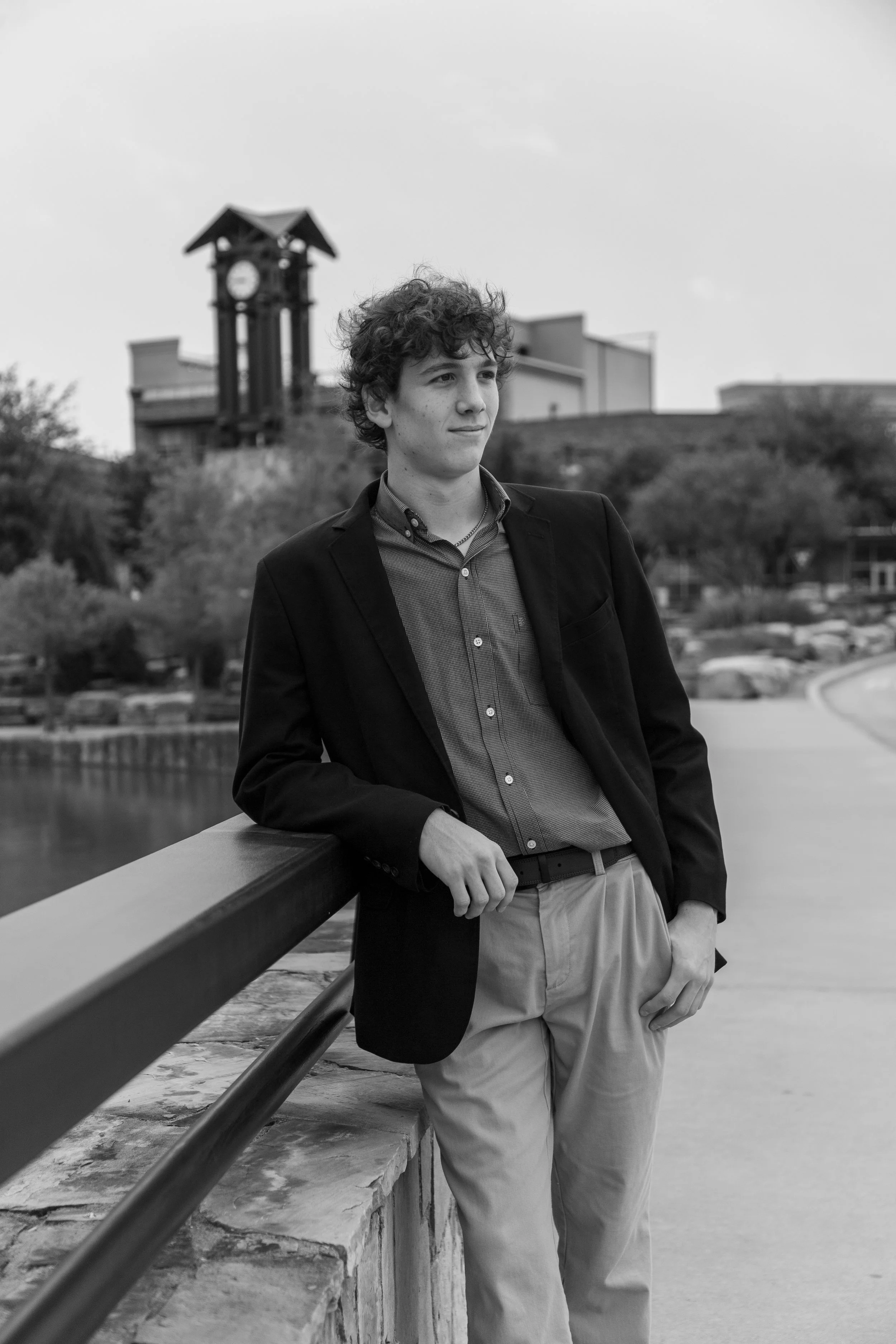 Black and white photo of a young man with curly hair wearing a blazer and shirt, leaning on a railing near a body of water, with trees and a building with a clock tower in the background.