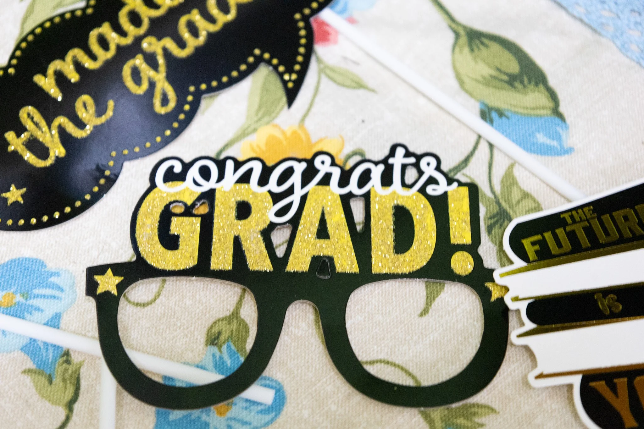 Celebration photo with decorative items reading "congrats GRAD!" and featuring glasses shape cutout, on a floral tablecloth, with additional grad-themed decorations.