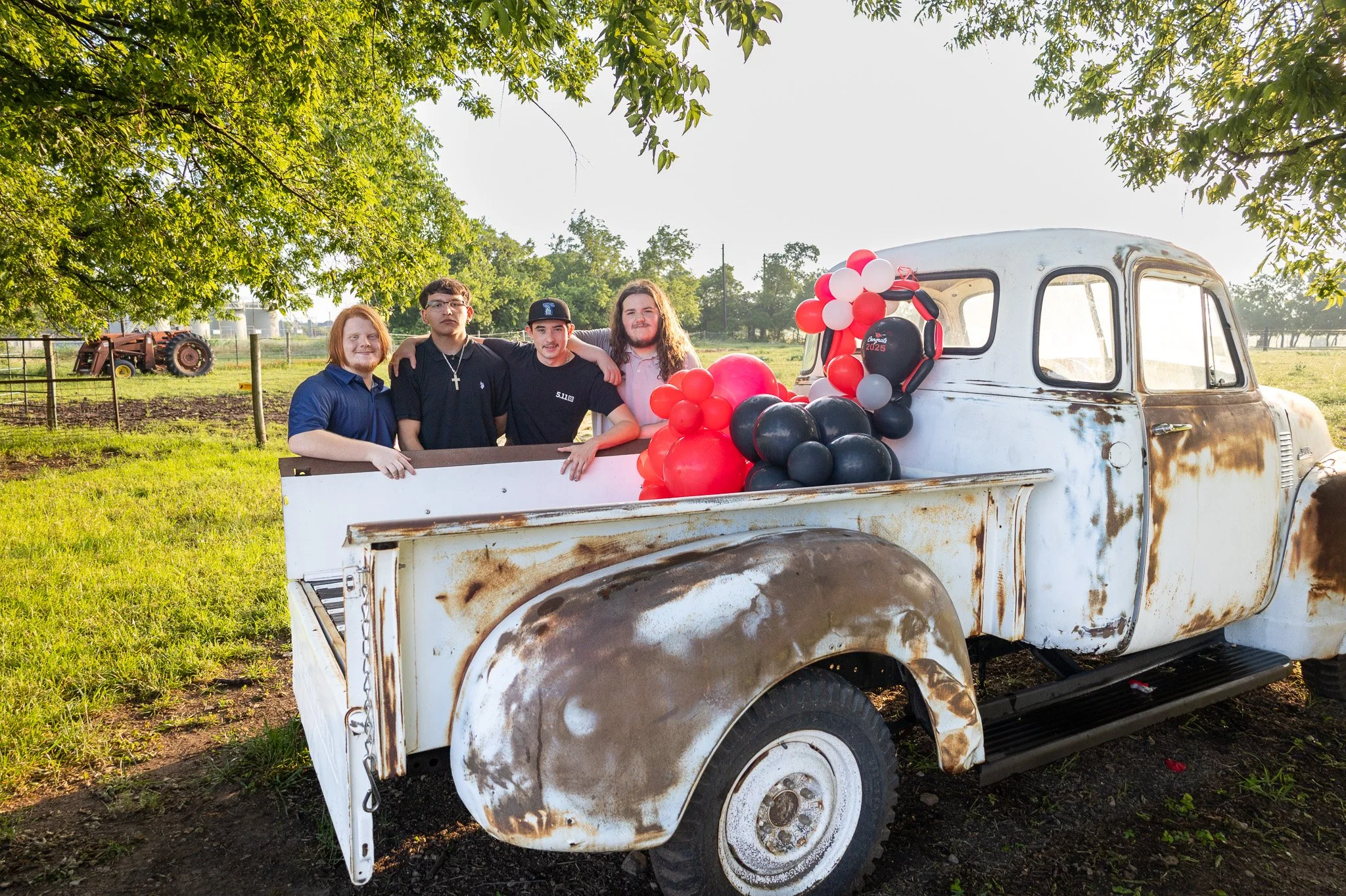 Four young men standing in the back of a white, rusty vintage pickup truck decorated with red, black, pink, and gray balloons, outdoors in a green field with trees and a tractor in the background.