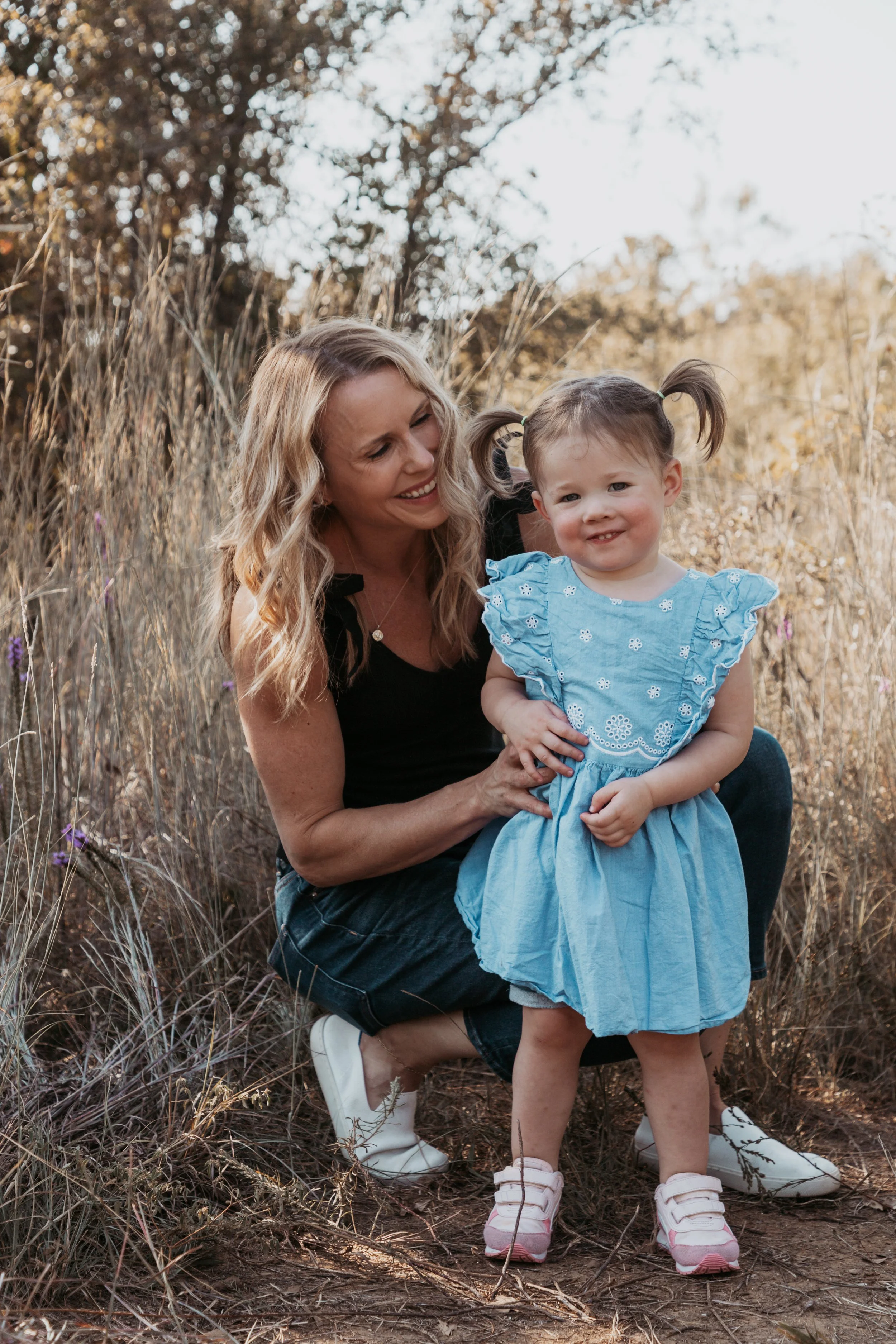 A woman and a young girl outdoors in a field of tall grass and wildflowers, smiling and enjoying a moment together.
