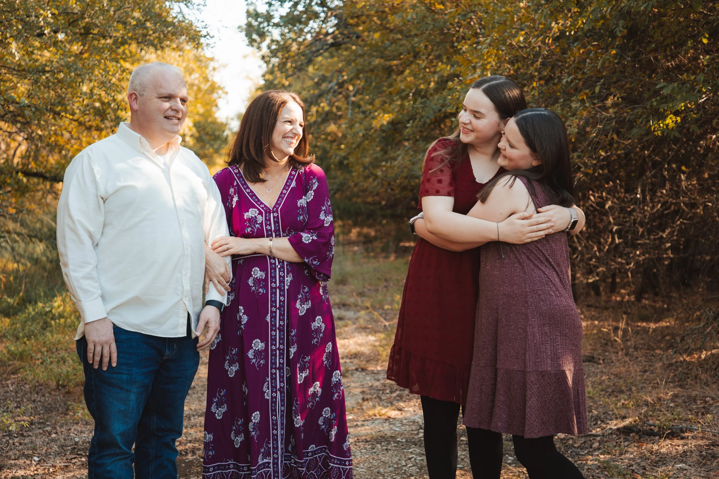 A family of five standing outdoors in autumn, smiling and hugging, with trees and fall foliage in the background.