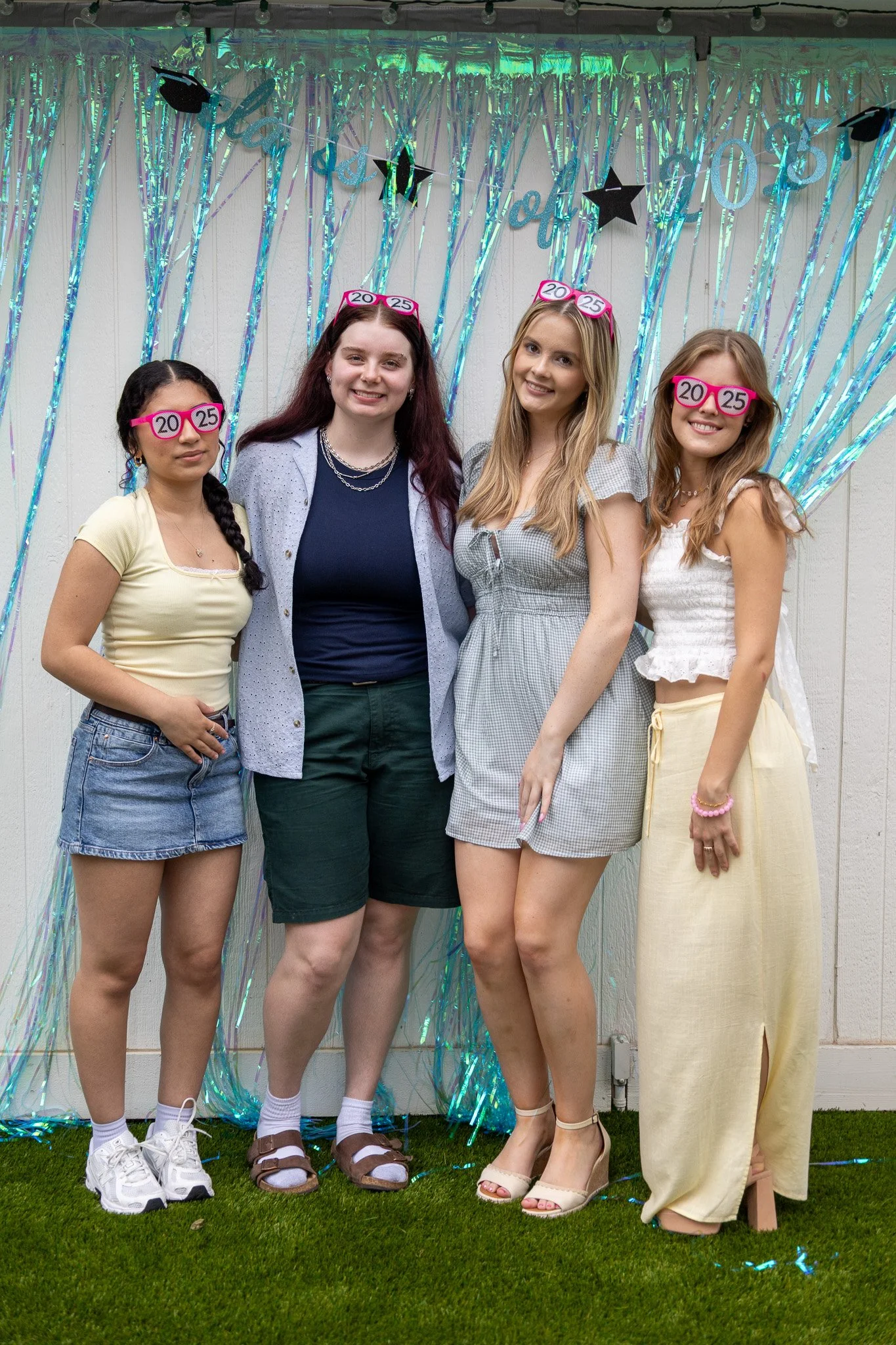 Four young women standing together at a graduation celebration, wearing silly glasses with '20' and '25' on them, in front of a shiny, decorative backdrop with hanging stars and a banner.