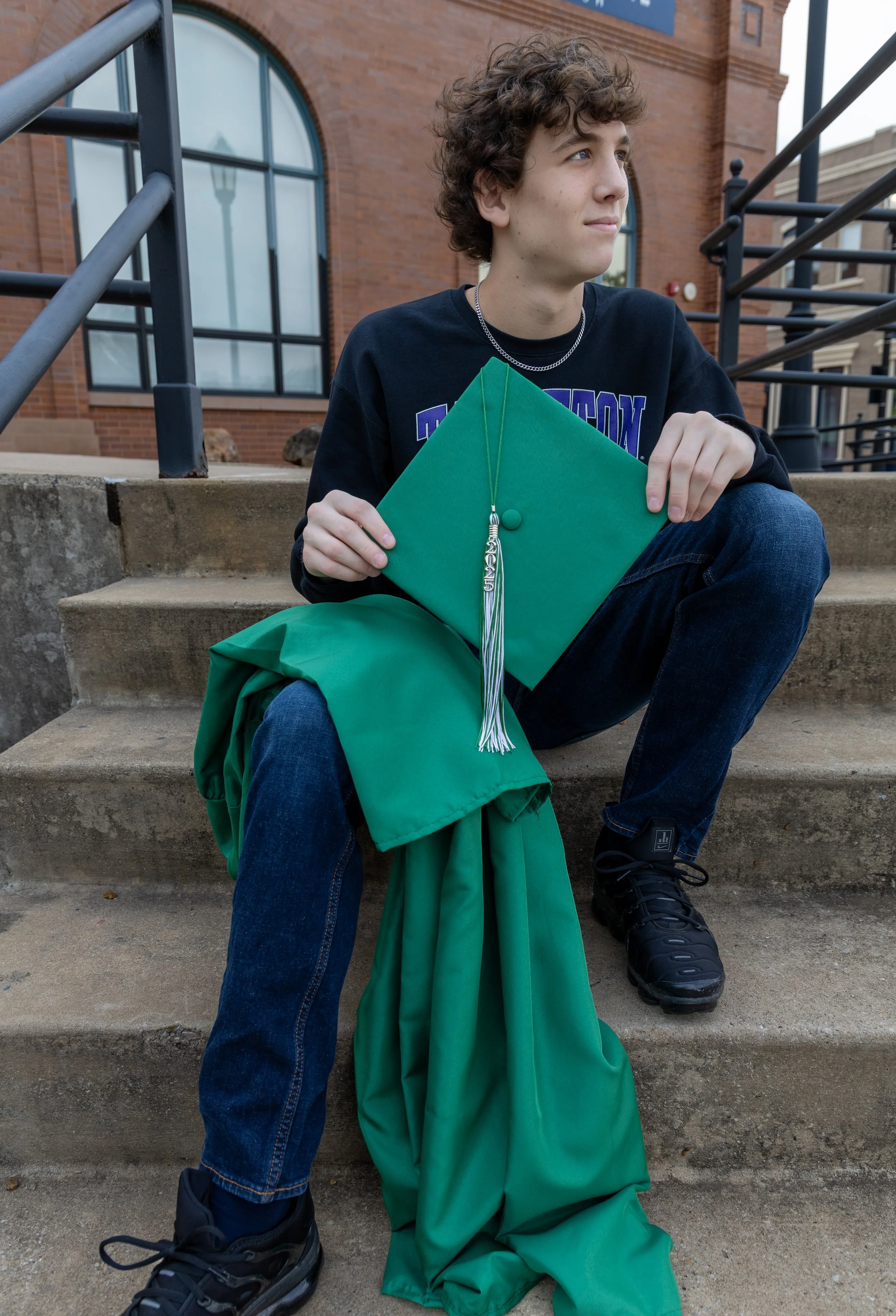A young man with curly hair sitting on outdoor concrete steps, holding a green graduation cap and a green gown, with a building featuring large arched windows in the background.