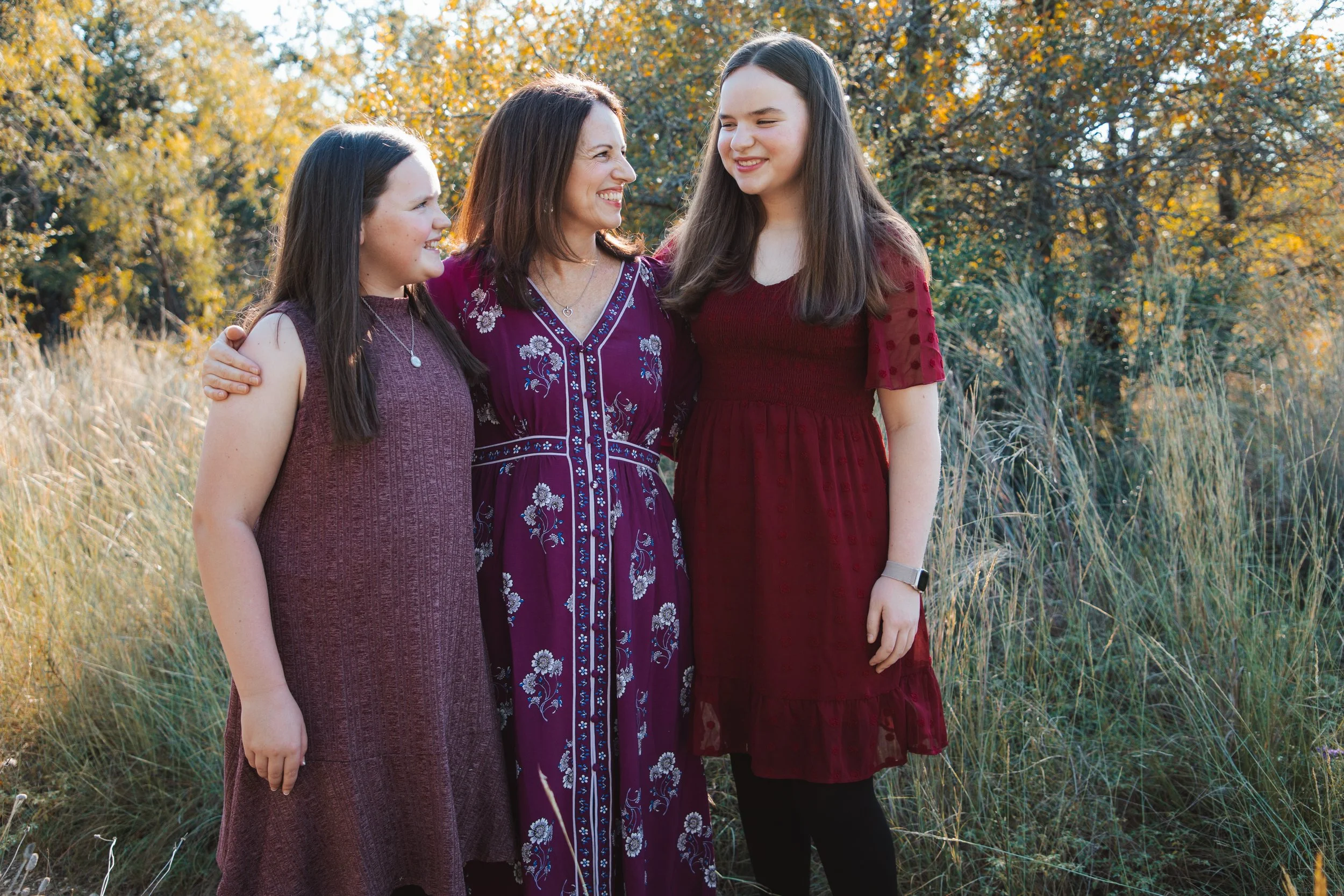 A woman and two girls standing outdoors in a field with autumn trees in the background. They are smiling and looking at each other.