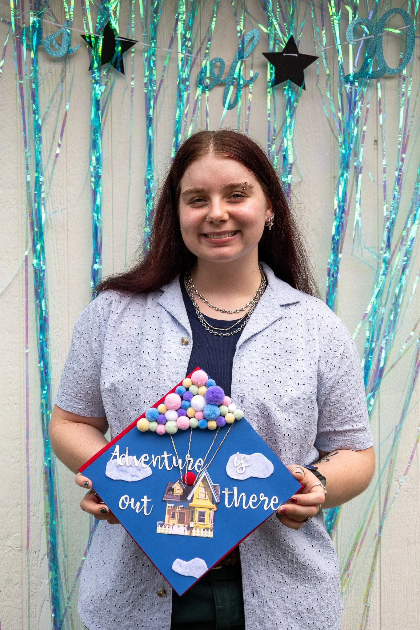 A young woman is standing in front of a decorated background with a graduation theme. She is smiling and holding a colorful craft sign that reads "Adventure is out there" with a house and clouds on it.