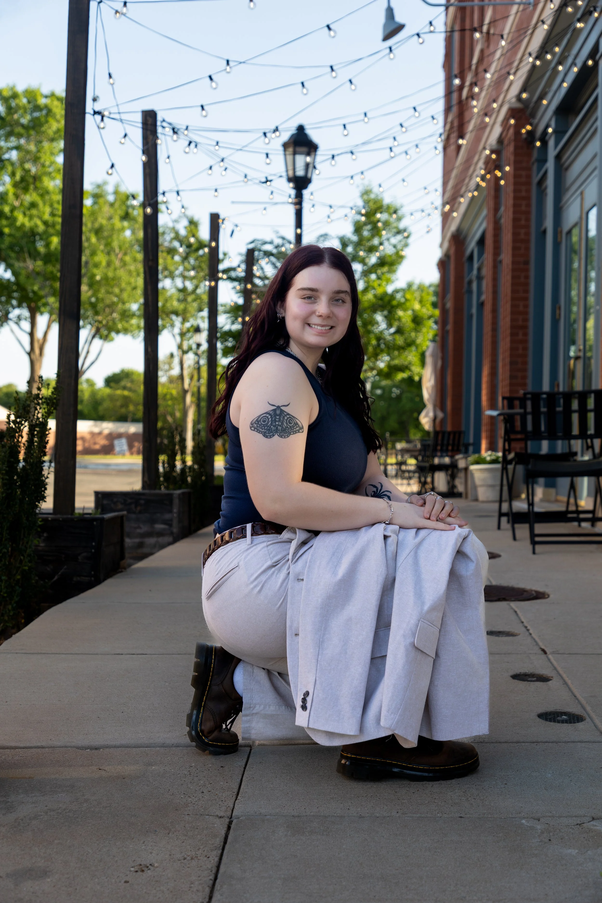 A young woman with dark hair, tattoos on her arms, wearing a black sleeveless top, beige pants, and black boots, is squatting on a sidewalk outdoors during daytime. She is smiling at the camera with string lights hanging overhead, green trees in the 