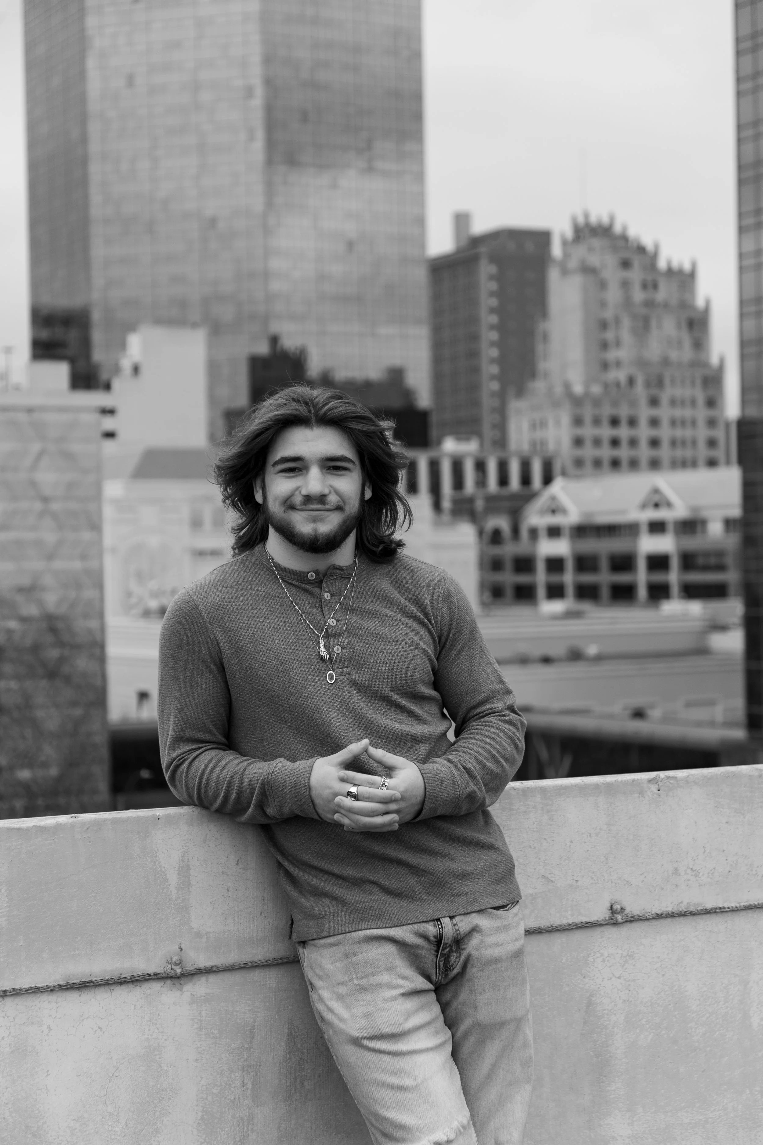 A young man with long hair and a beard smiling while standing on a rooftop with city skyscrapers in the background, in black and white.