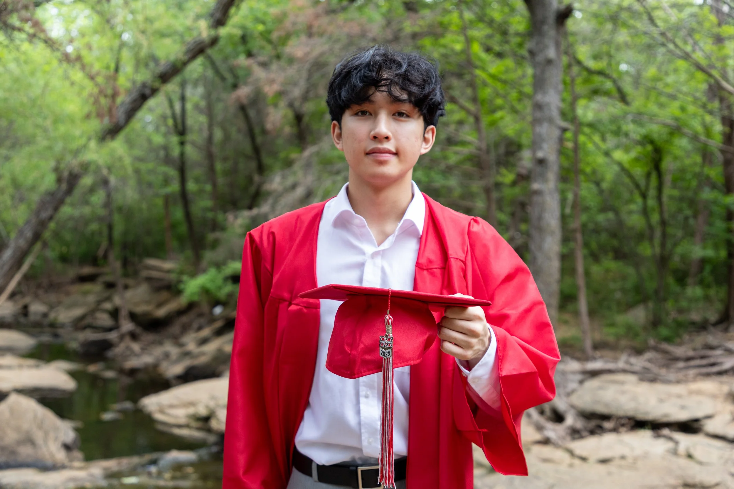 Young man in a red graduation gown holding a red cap outdoors in a wooded area.