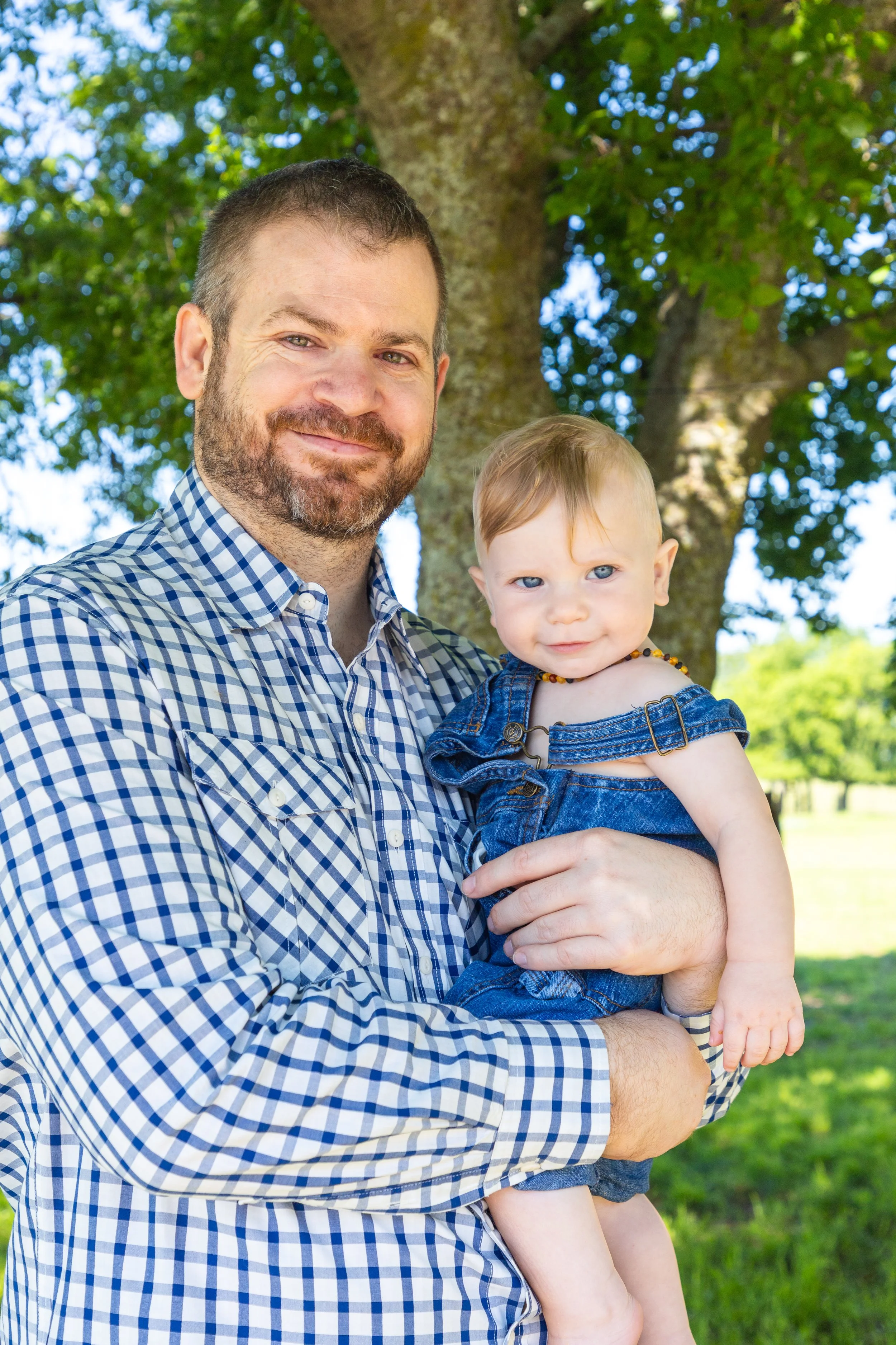 A man holding a young girl outdoors in front of a large tree with green leaves, smiling at the camera.