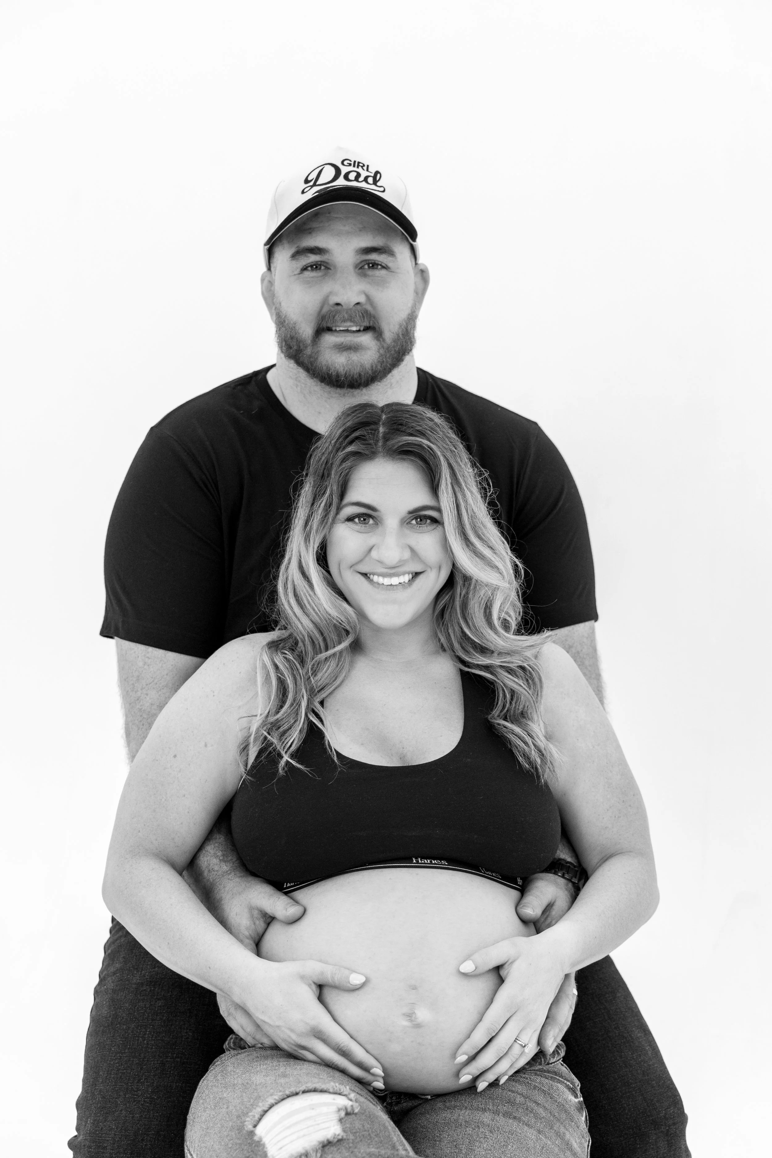 A pregnant woman seated and smiling, with a man behind her embracing her, both appearing happy against a plain white background.