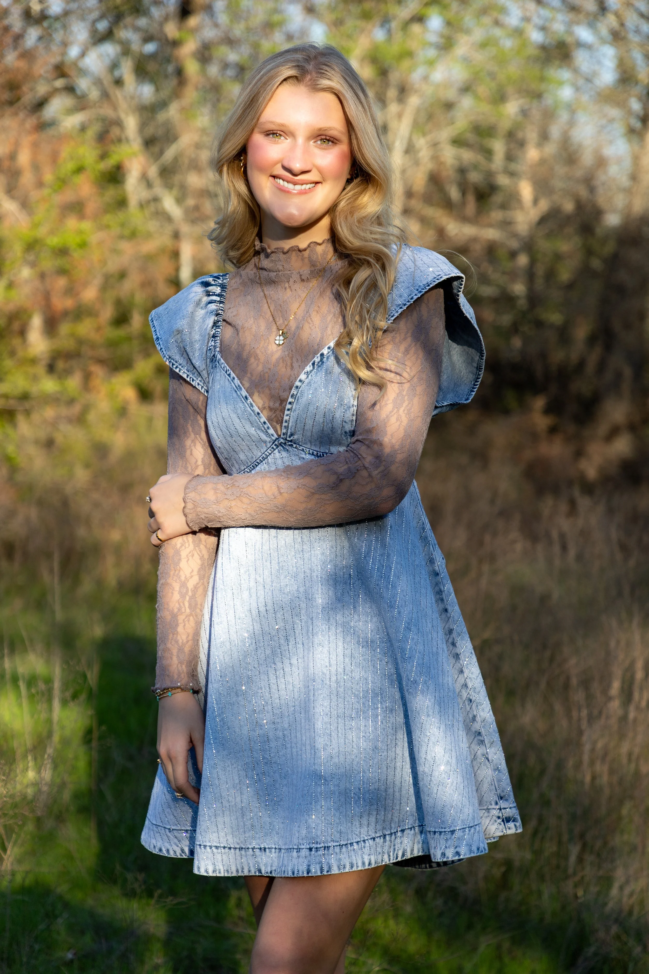 Young woman with blonde wavy hair smiling outdoors, wearing a black lace long-sleeve top with a collared denim sleeveless dress over it, with trees and grass in the background.