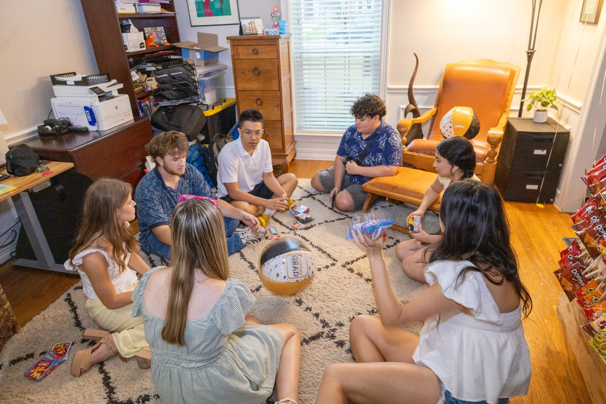 Group of young people sitting on a rug playing cards and chatting in a cozy living room with wooden furniture and snacks.
