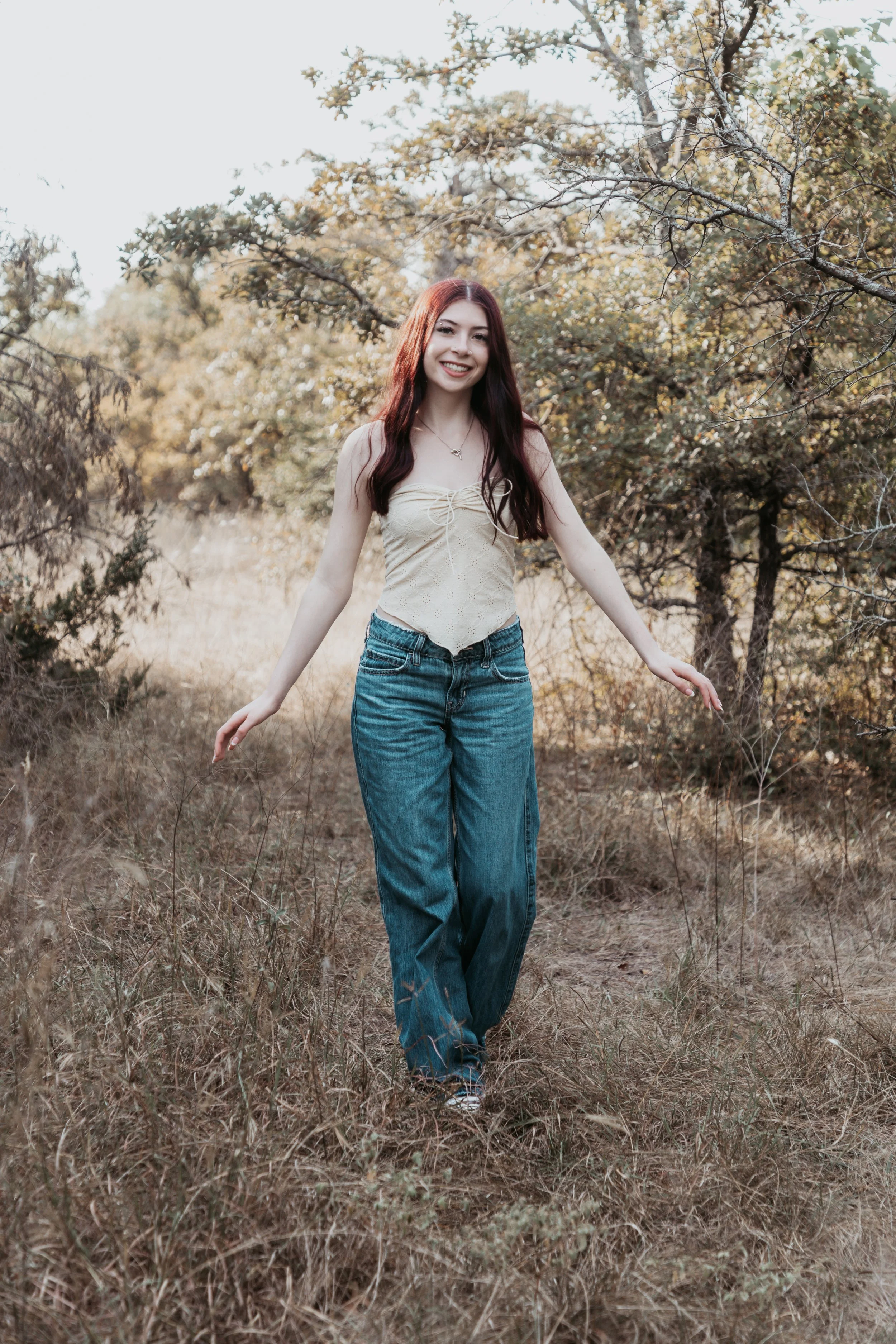 A young woman with long red hair smiling and walking in a grassy outdoor area with trees and foliage in the background.