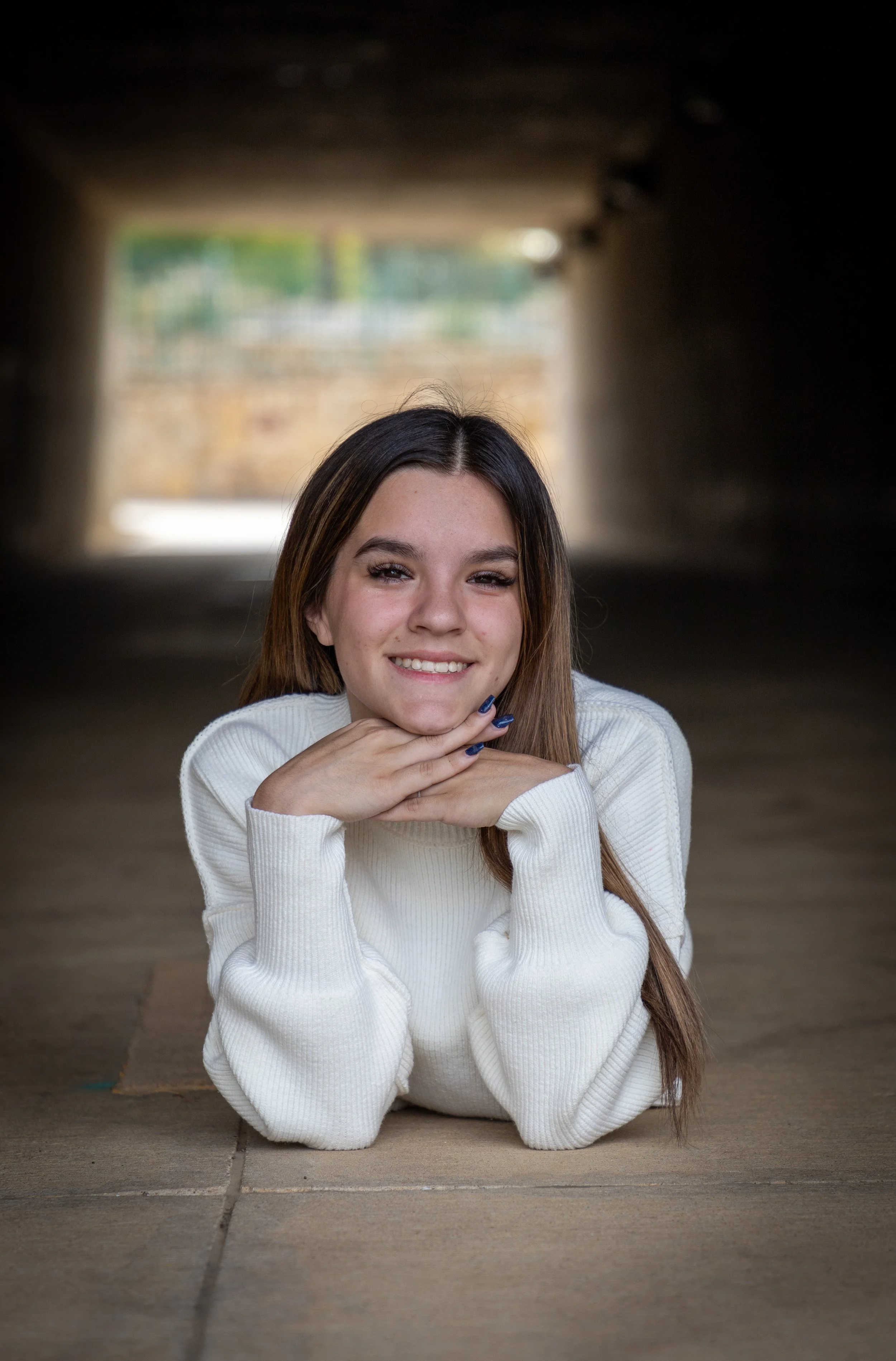 Young woman with long brown hair wearing a white sweater, lying on the ground under a bridge or tunnel, smiling with her chin resting on her hands.