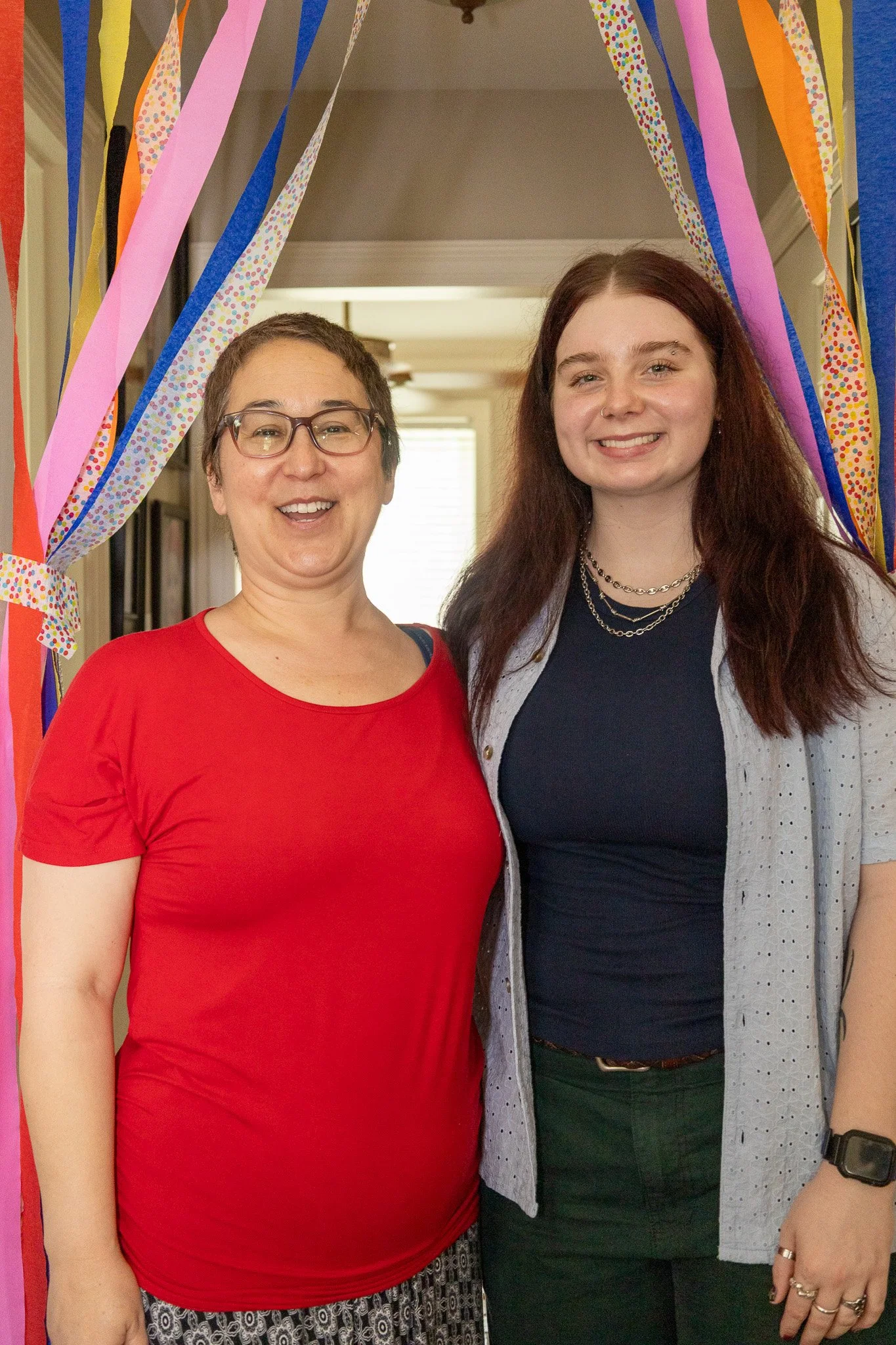 Two women smiling and standing under colorful streamers at a celebration.