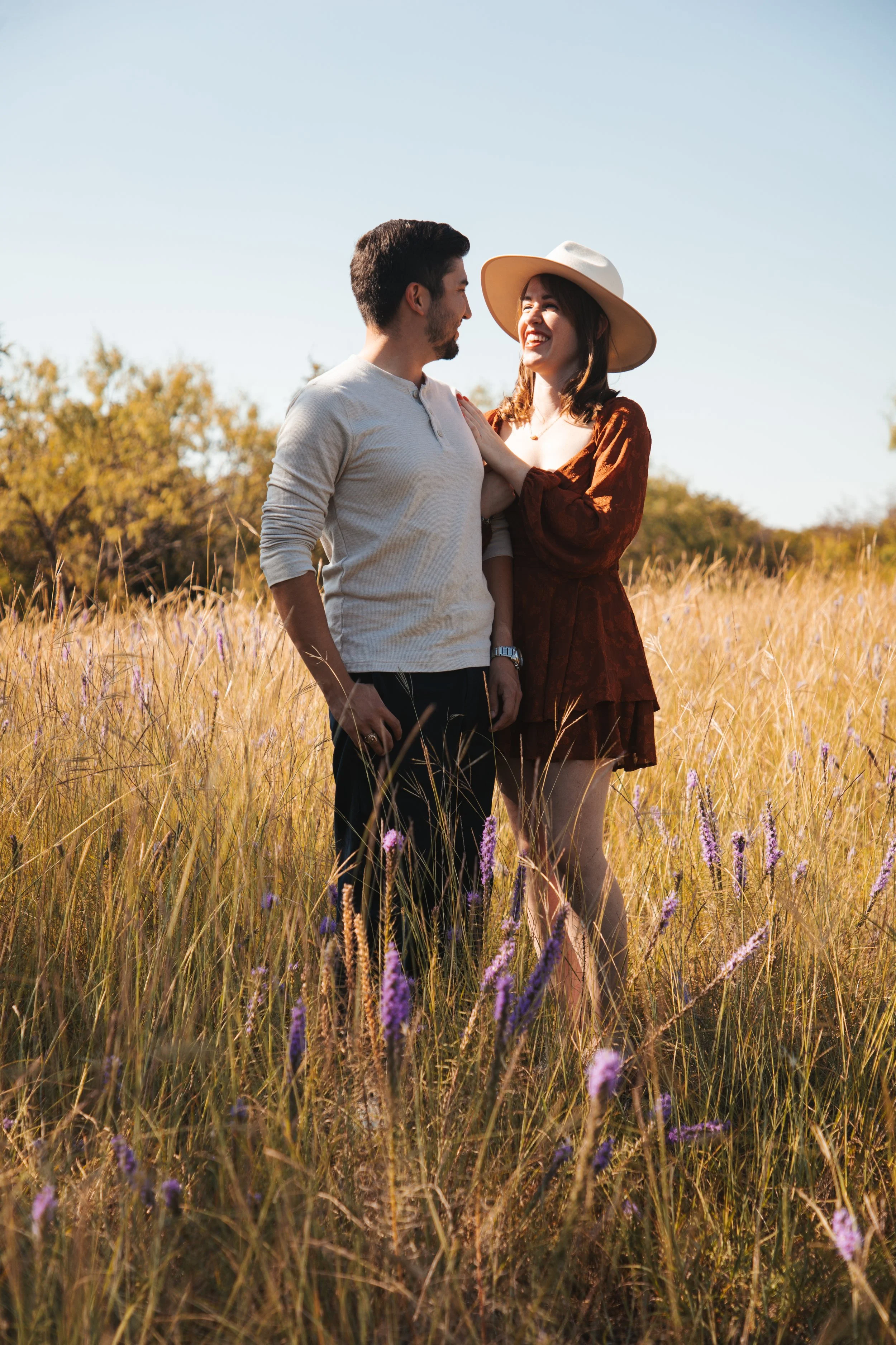 A couple standing in a field of tall grass and purple flowers, smiling at each other on a sunny day.